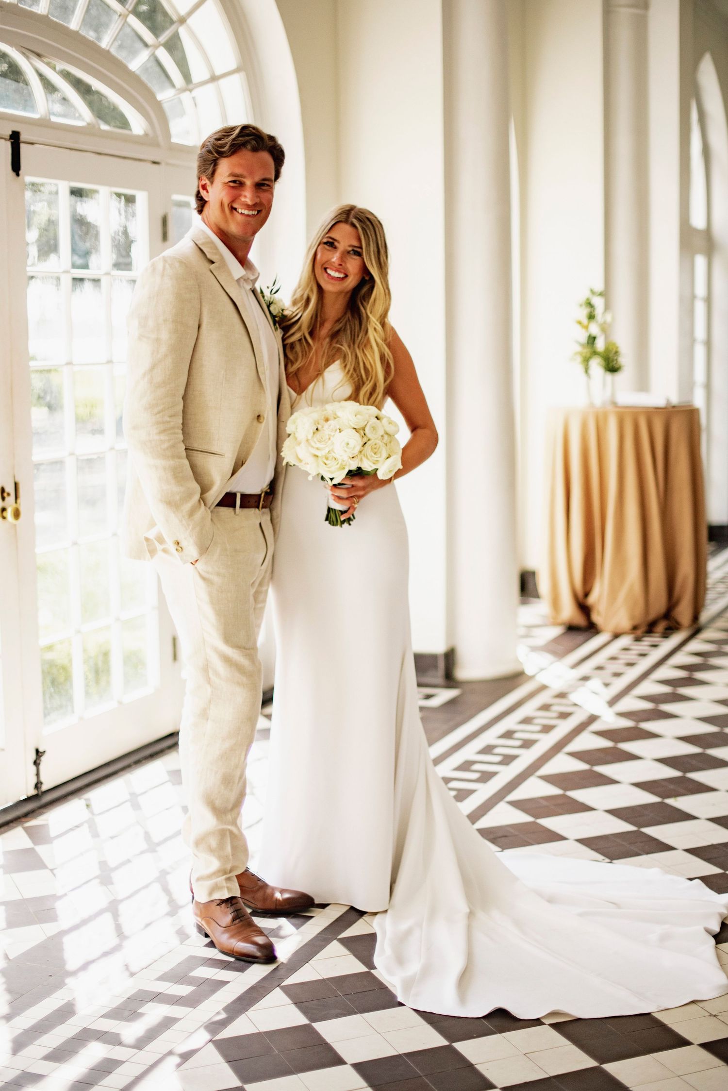 A couple in wedding attire poses on a black and white checkered floor in an elegant sunlit hallway.