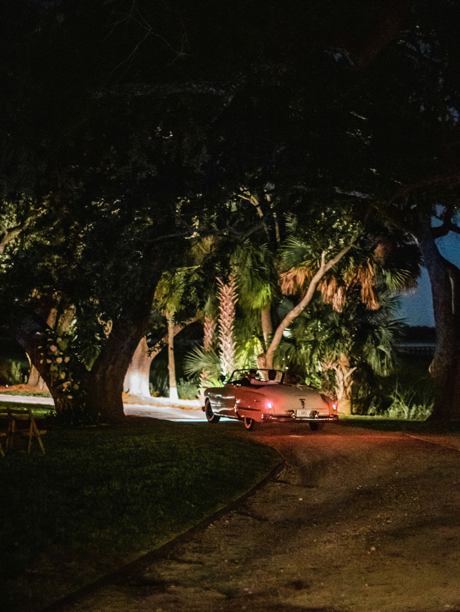A vintage red car parked under illuminated palm trees at night on a dirt path.
