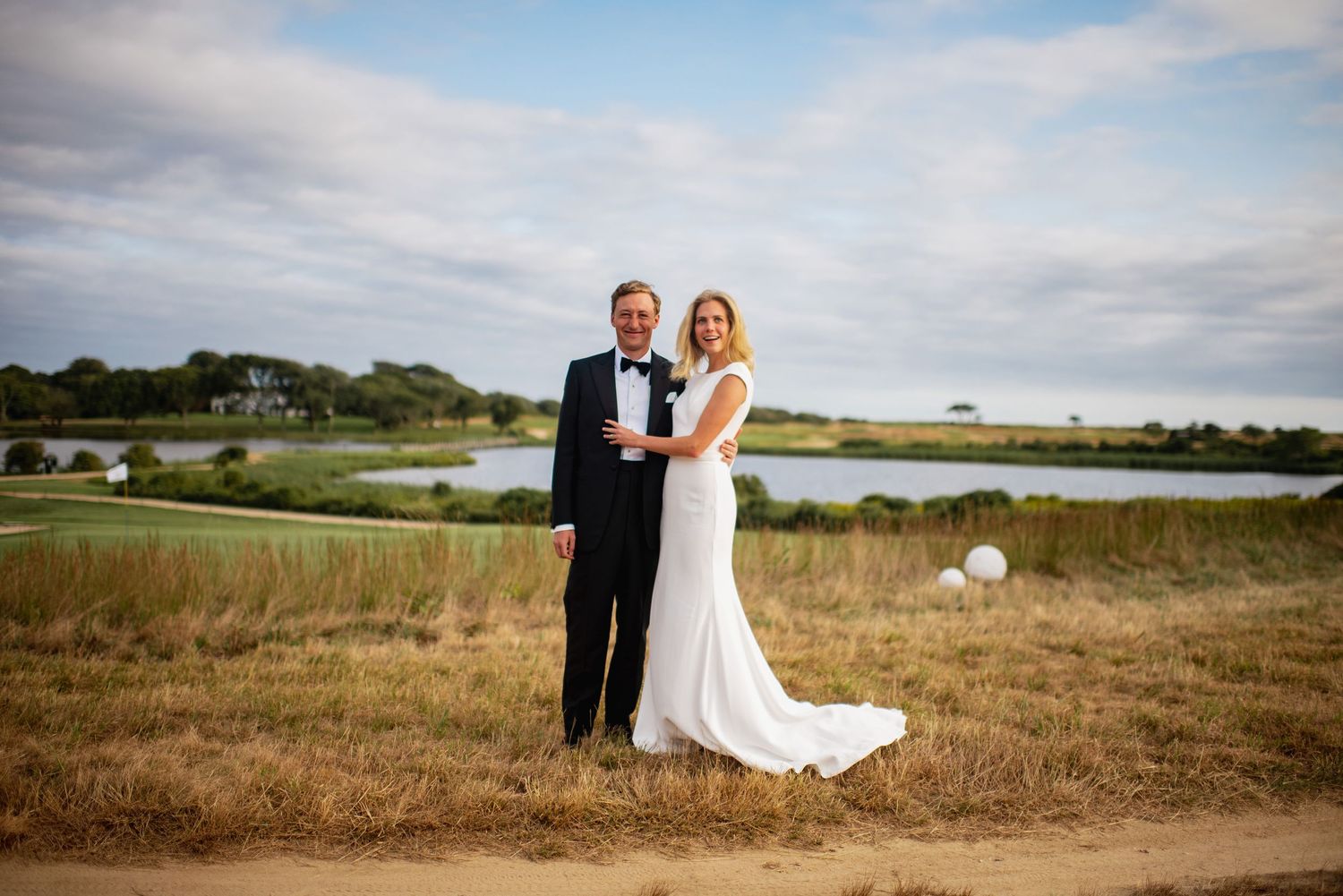 A couple in formal wedding attire poses together in a scenic coastal landscape at sunset.