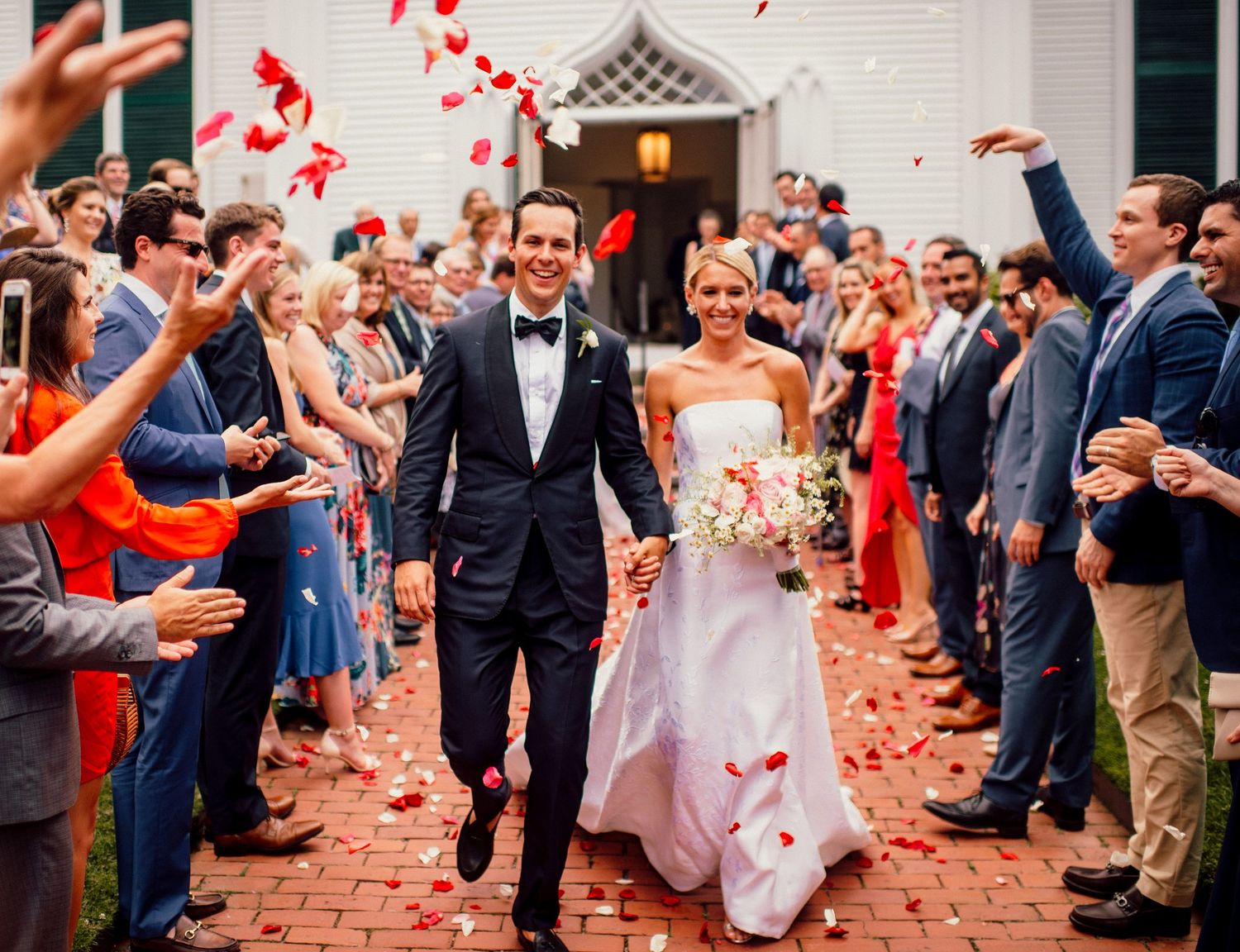 Wedding guests throw red rose petals as newlyweds run through brick pathway.