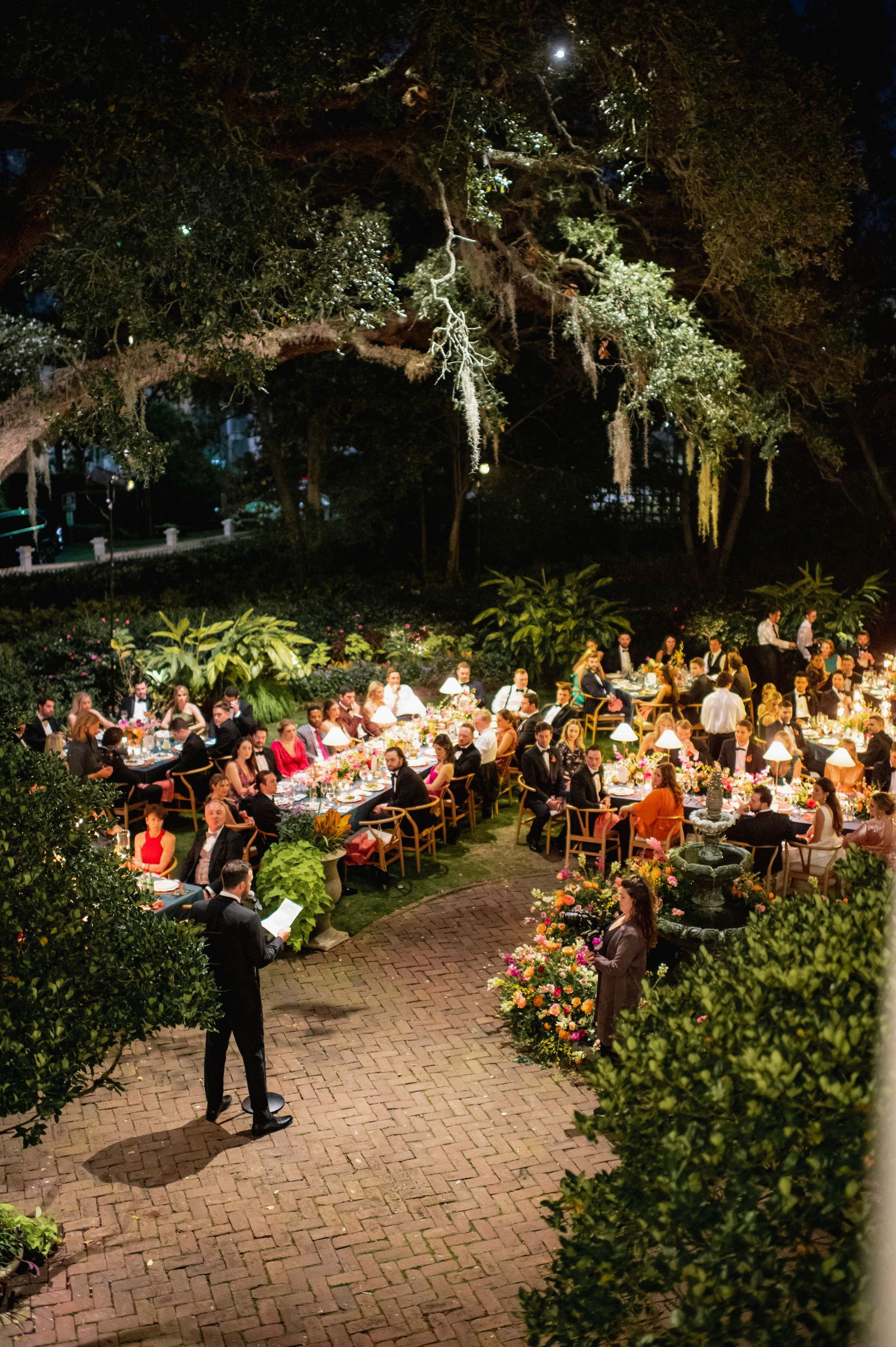 Outdoor evening reception with guests seated at illuminated tables along a brick pathway.