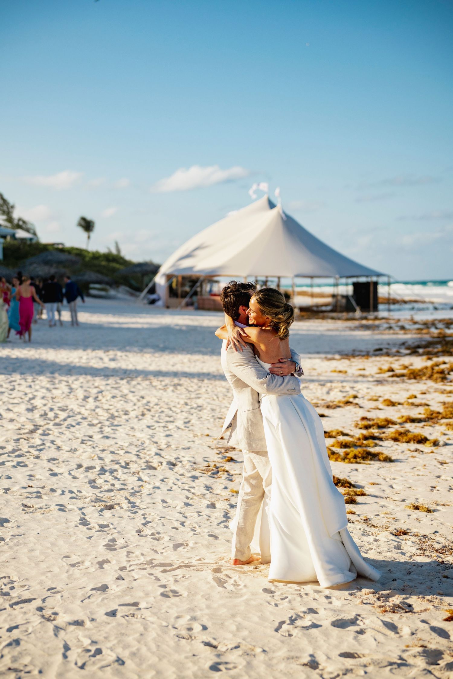 Couple embracing on a white sandy beach with event tents in the background.
