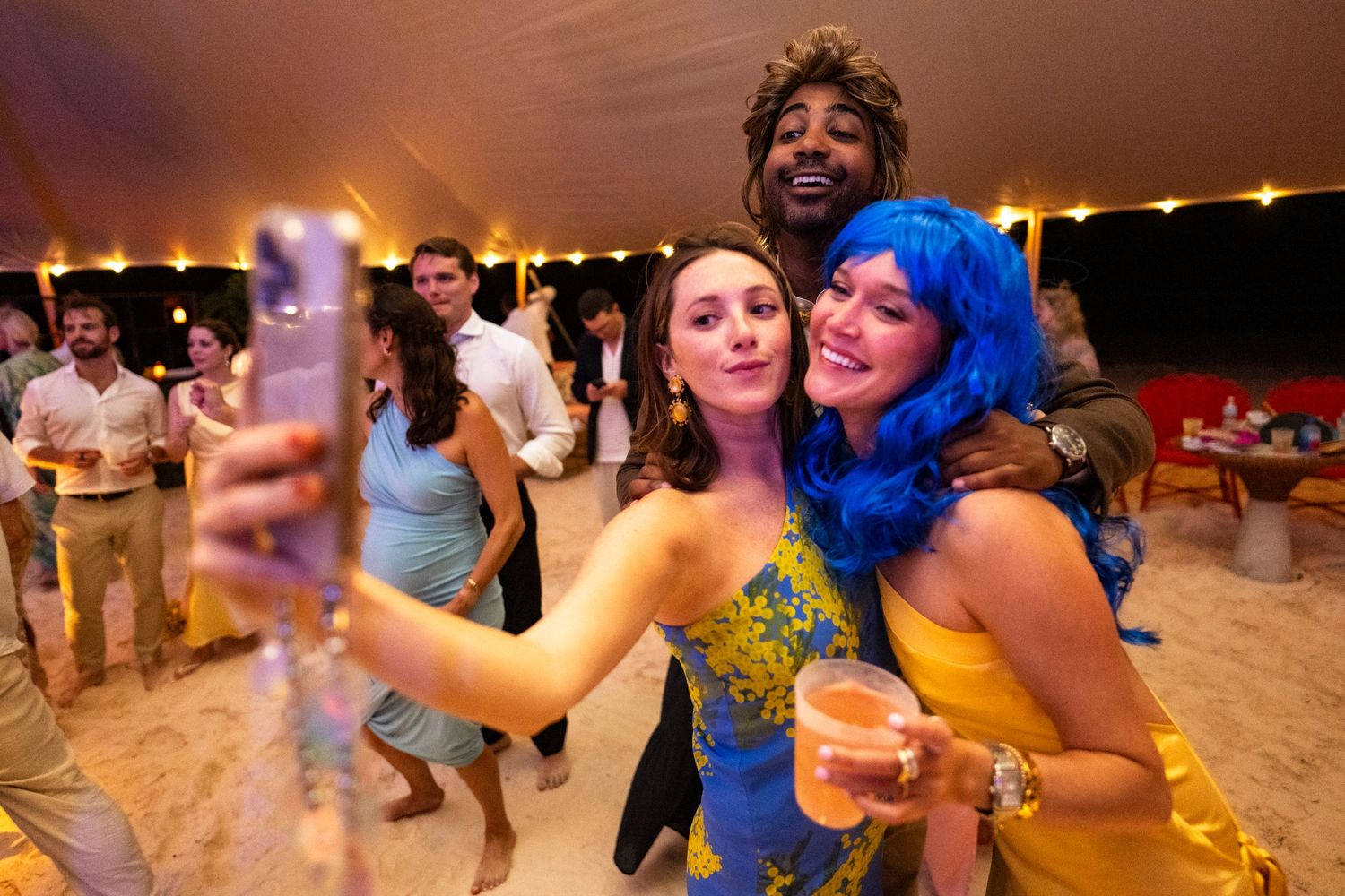 Party guests taking selfies and dancing under tent at beach celebration.