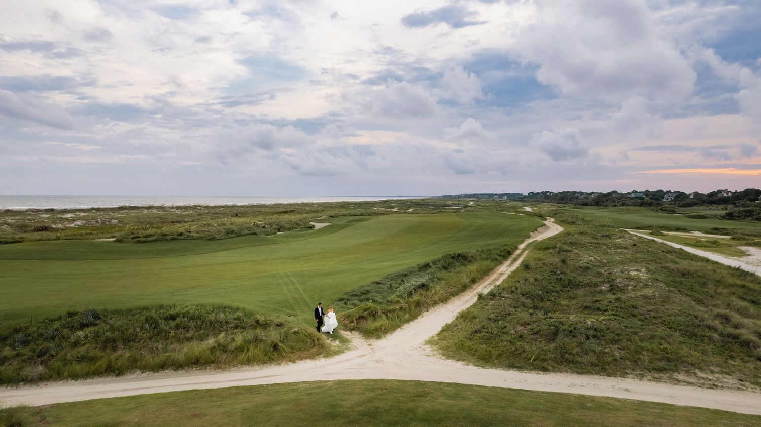 A panoramic view of a scenic golf course with rolling green hills, winding paths and cloudy skies above.