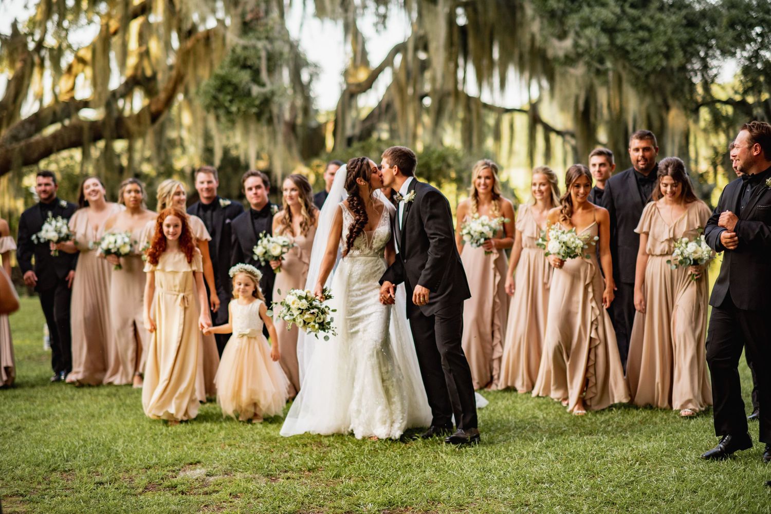 Wedding party in neutral champagne dresses and dark suits poses on a grassy lawn under Spanish moss trees.