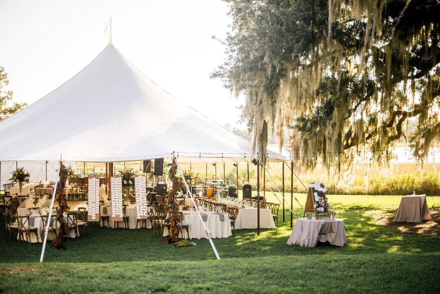 Elegant outdoor wedding reception setup with white tent, hanging decor and Spanish moss draped trees at sunset.