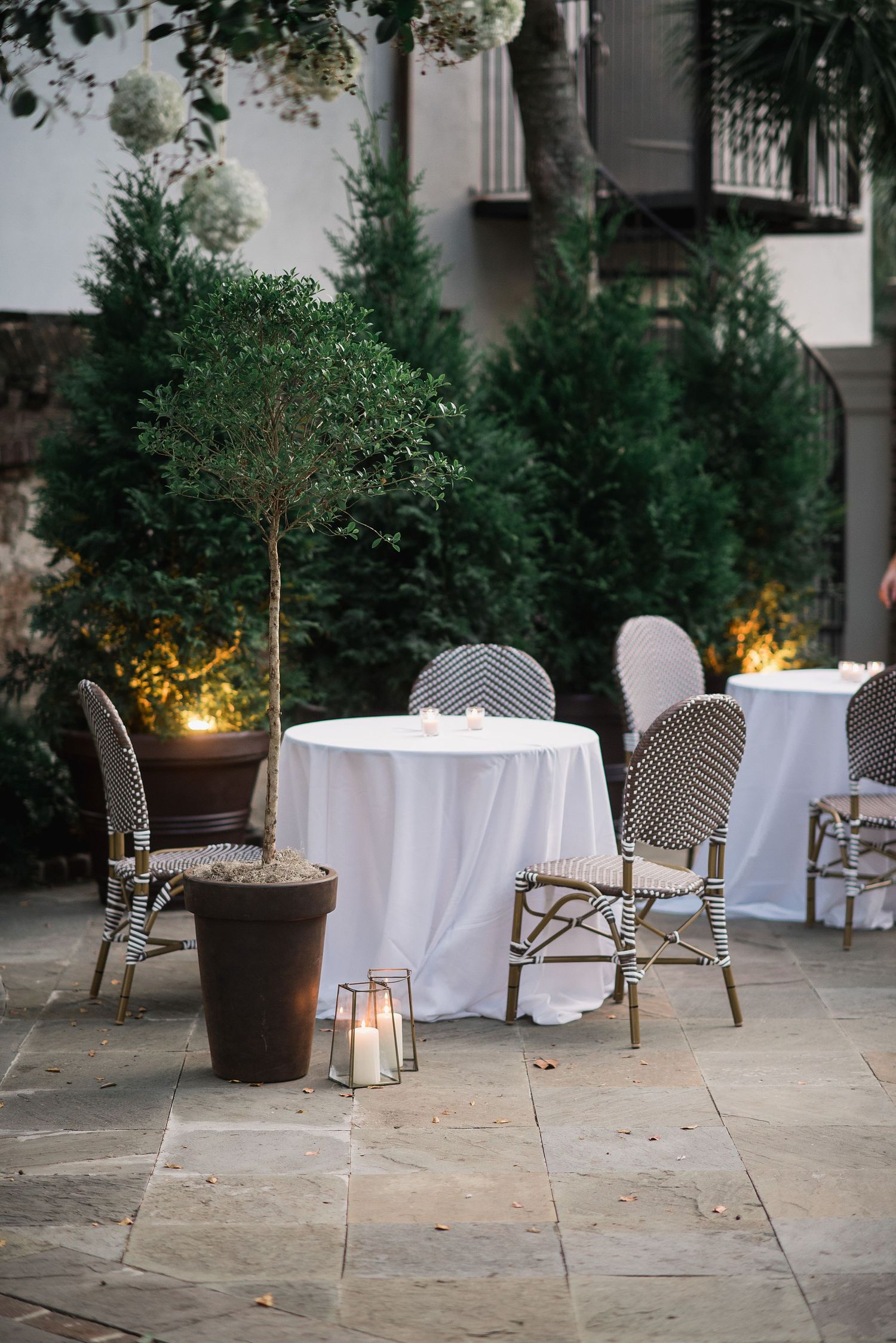 Outdoor dining setup with white tablecloths, bistro chairs and potted evergreen trees illuminated by soft candlelight.