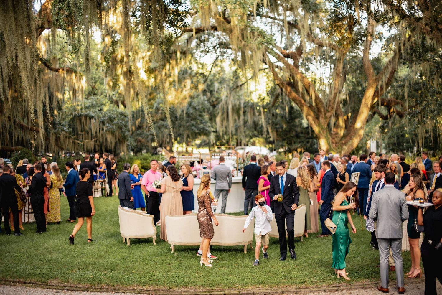 Outdoor wedding reception under Spanish moss-draped oak trees with guests mingling on a lush lawn at sunset.