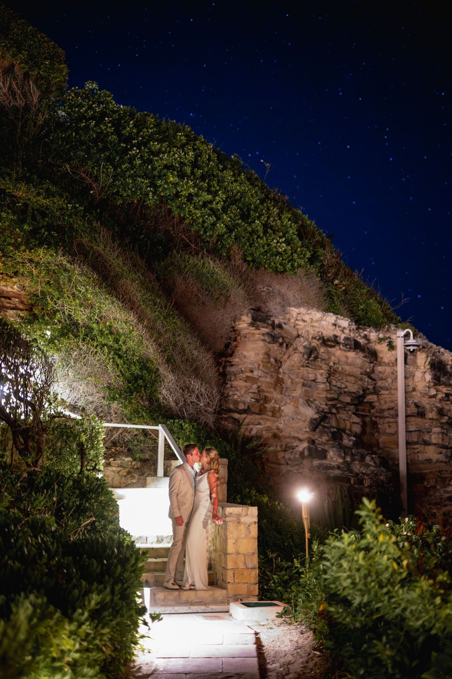 Outdoor basketball court illuminated at night with rocky cliff wall and foliage visible in the background.