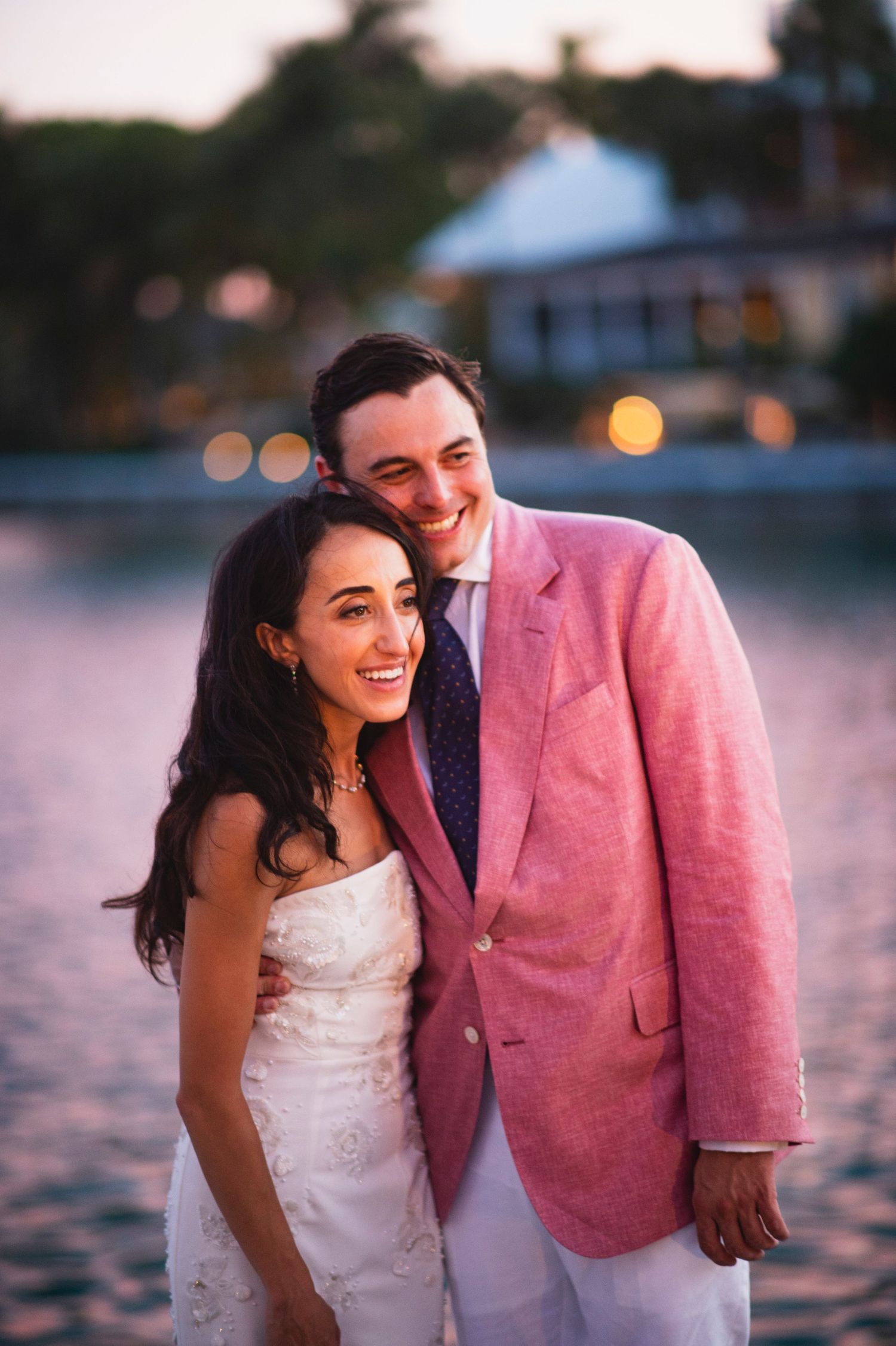 A couple shares a joyful moment at sunset by the water, with the person wearing a pink blazer and white dress.