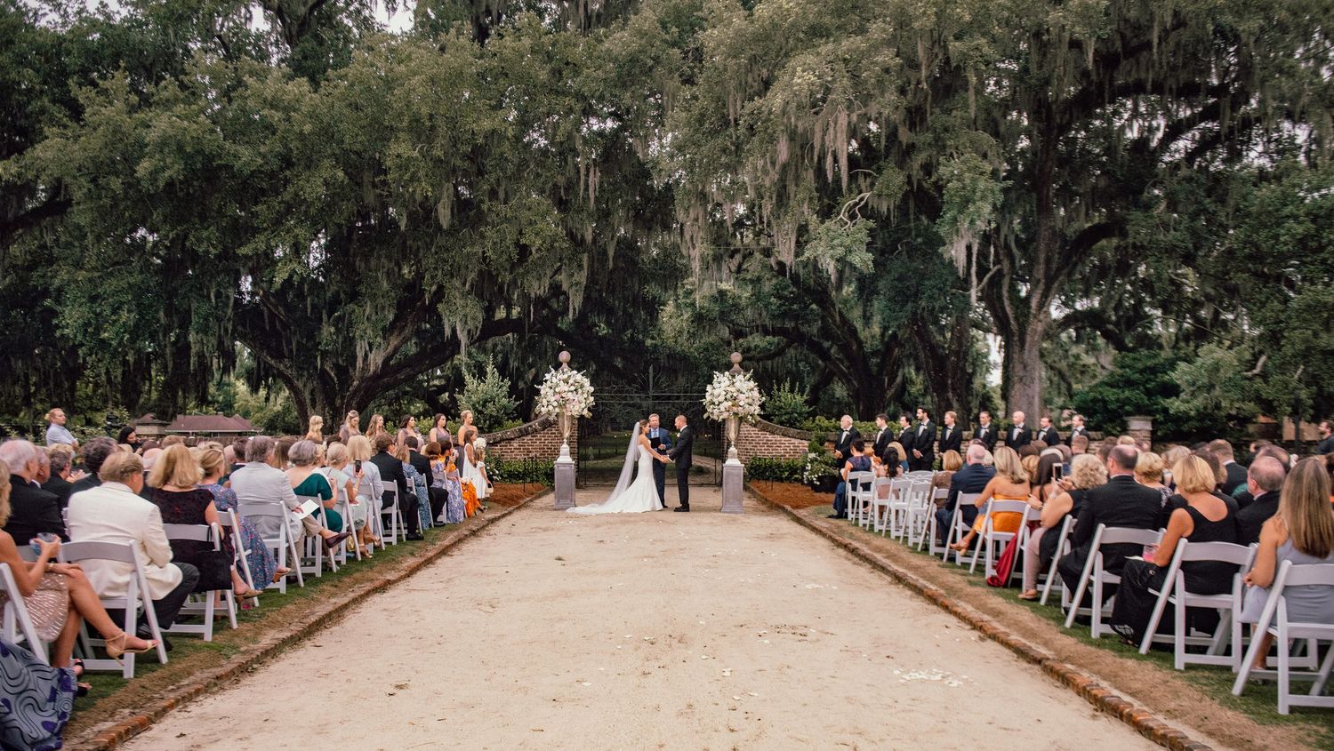 Outdoor wedding ceremony setup with white chairs arranged in rows along a tree-lined path leading to a decorated altar.