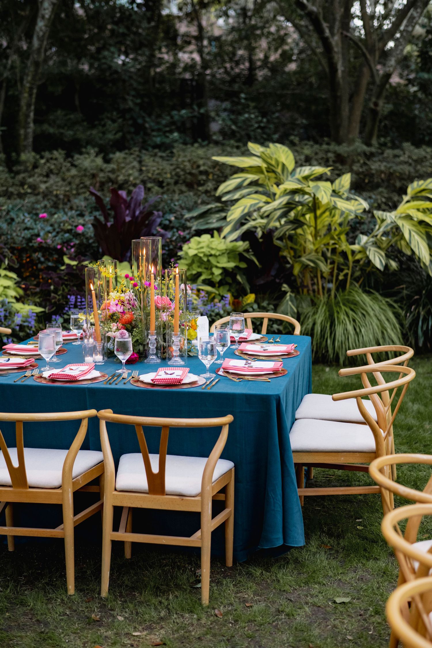 Outdoor garden party table setting with teak chairs and blue tablecloth decorated with colorful floral centerpieces.