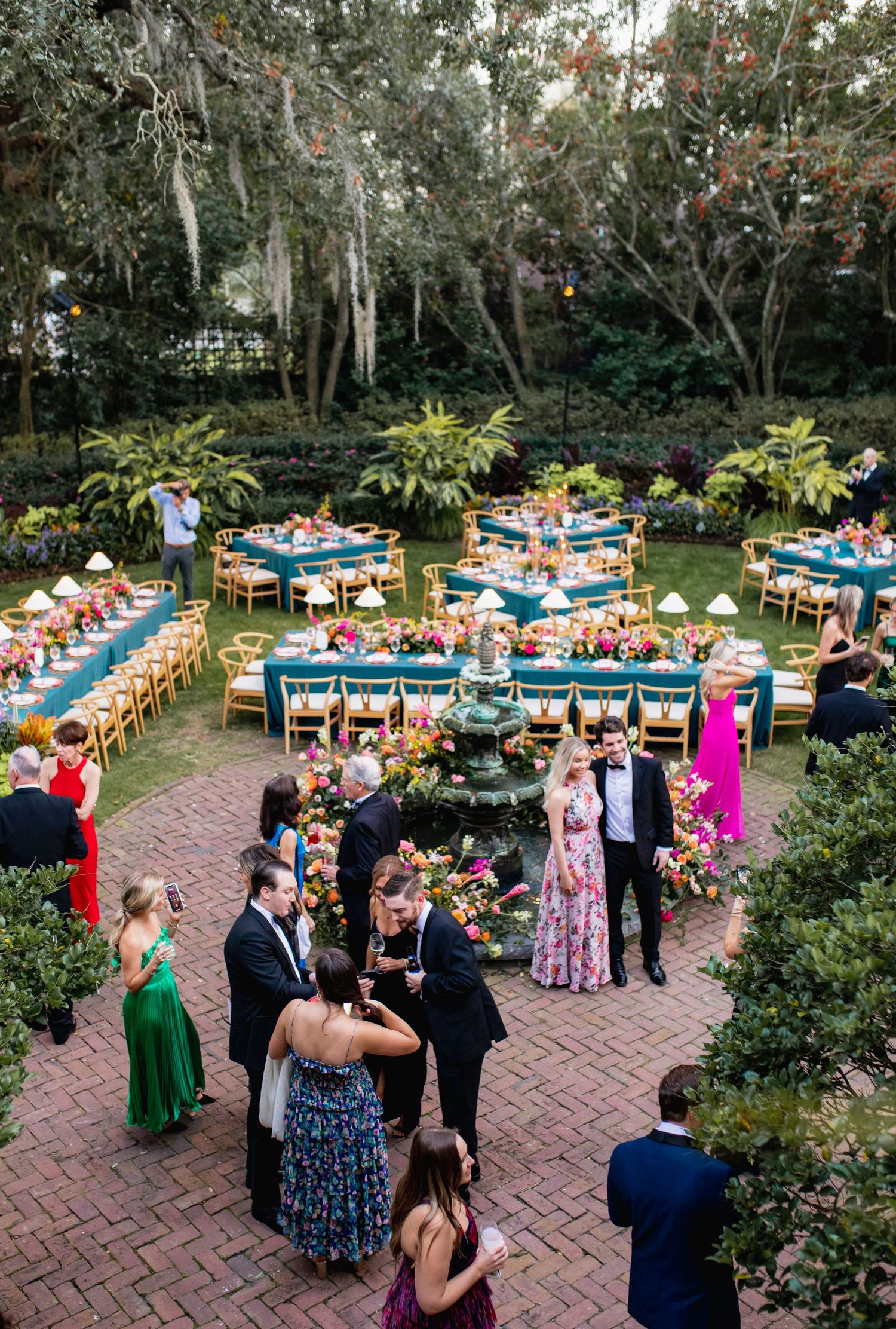 Guests mingle during an outdoor garden reception with long tables decorated in blue and orange under tree canopies.