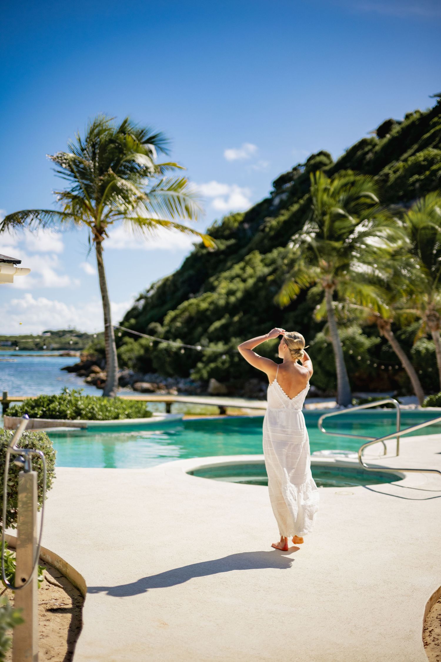 Woman in white dress walks by tropical pool with palm trees and ocean view.