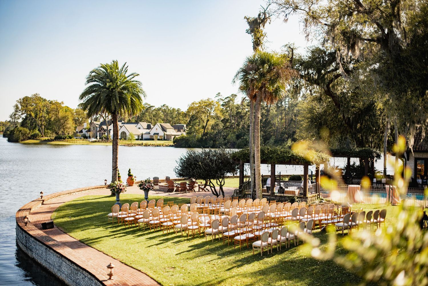 Outdoor waterfront wedding venue with rows of white chairs set up on manicured lawn beside palm trees and lake.