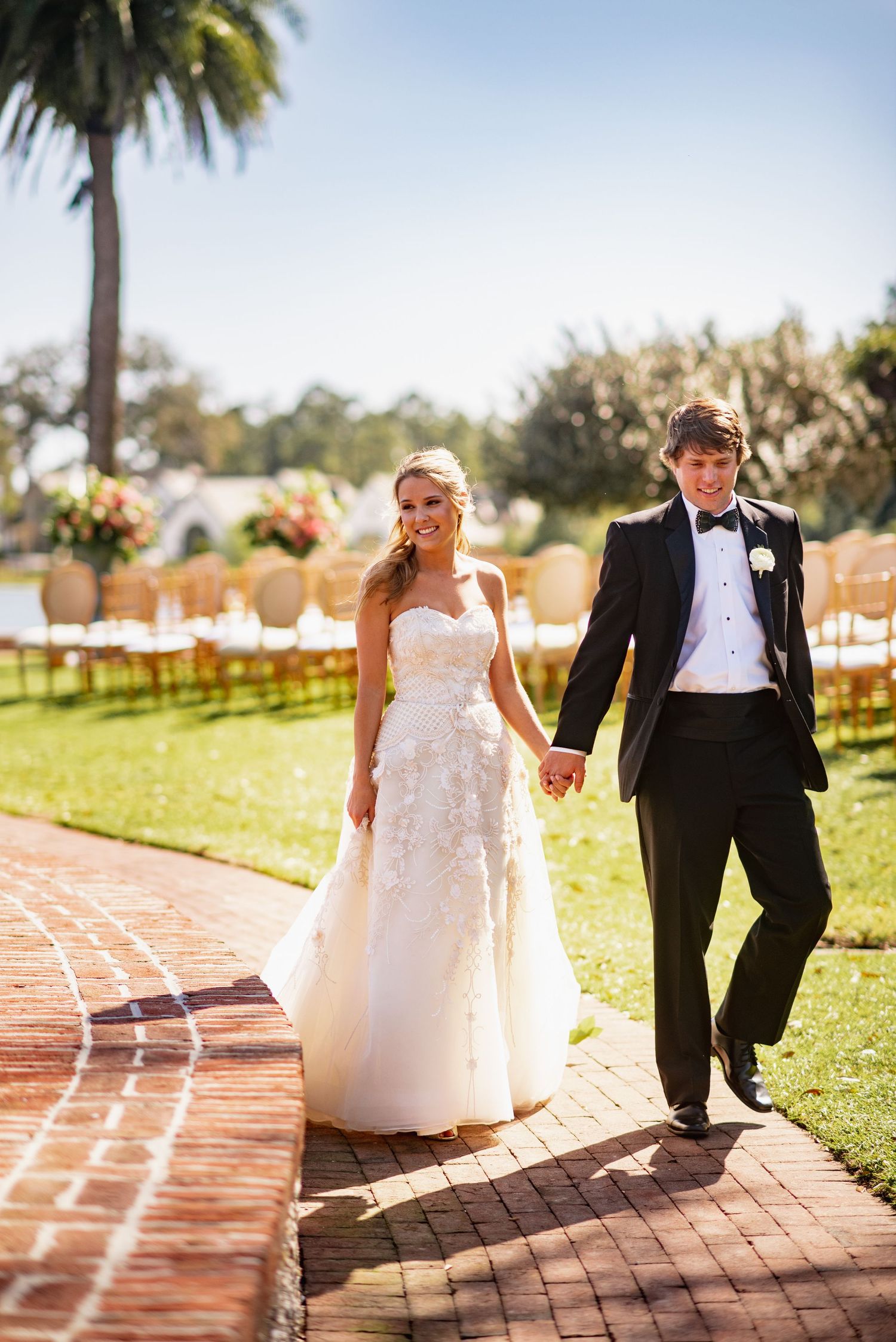 A bride and groom walk hand in hand along a brick pathway under palm trees at an outdoor wedding venue.