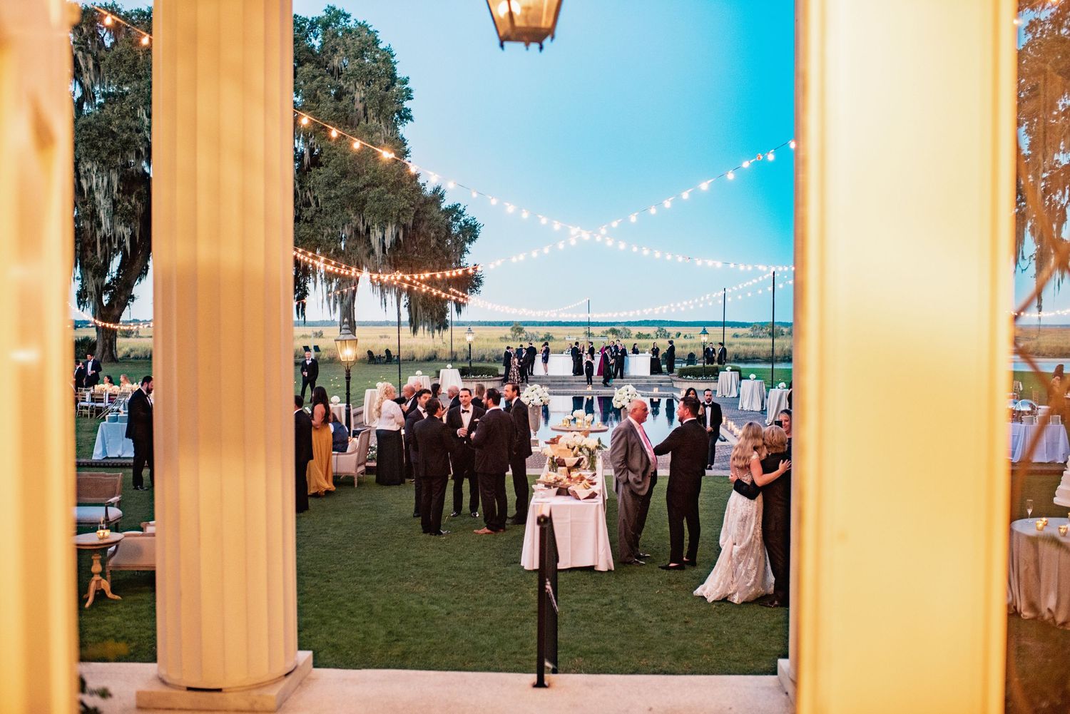Elegant outdoor wedding reception at dusk with string lights and columns framing guests gathered on a waterfront lawn.
