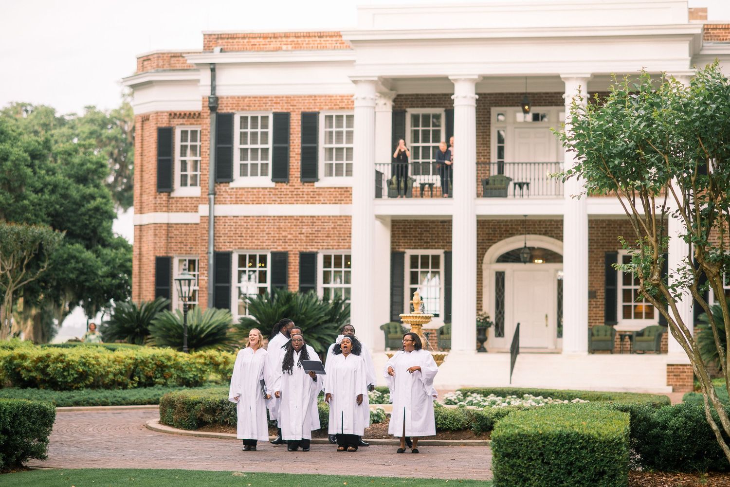 Graduates in white robes walk across a scenic college campus with brick buildings and manicured gardens.