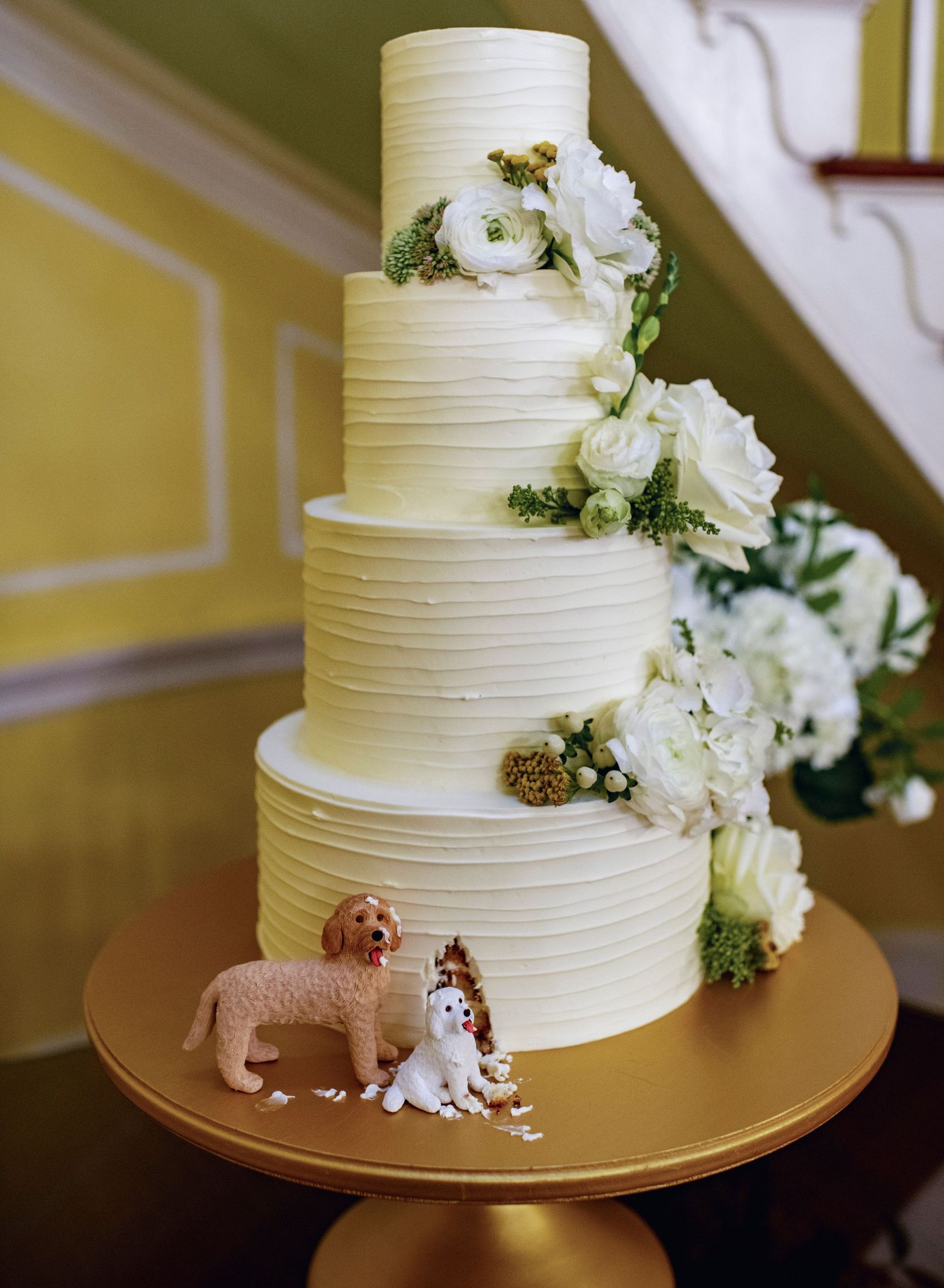 A white tiered wedding cake decorated with textured horizontal lines and fresh white flowers on a gold cake stand.