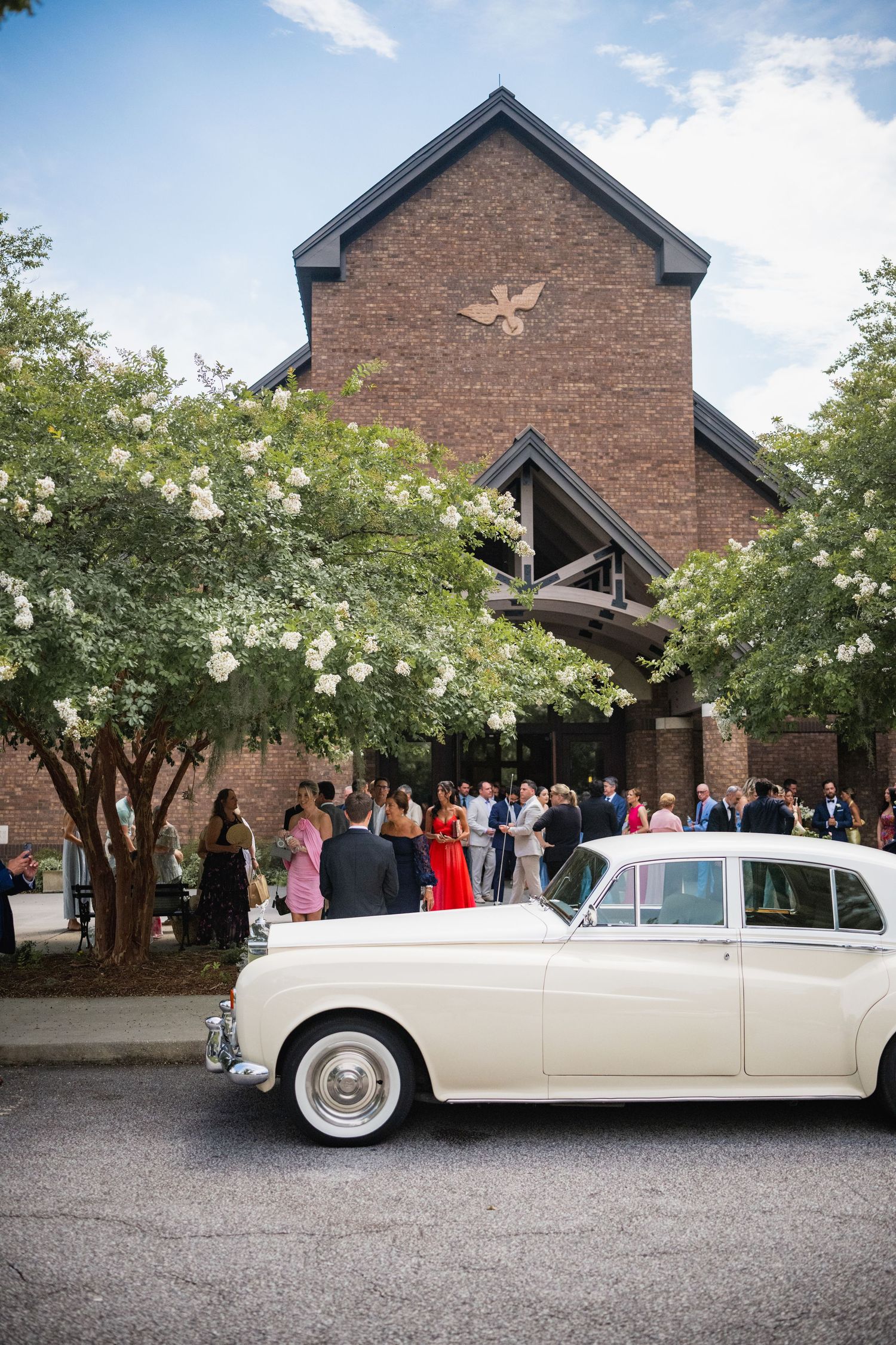 Classic white vintage car parked outside brick church building with flowering trees and wedding guests gathered.