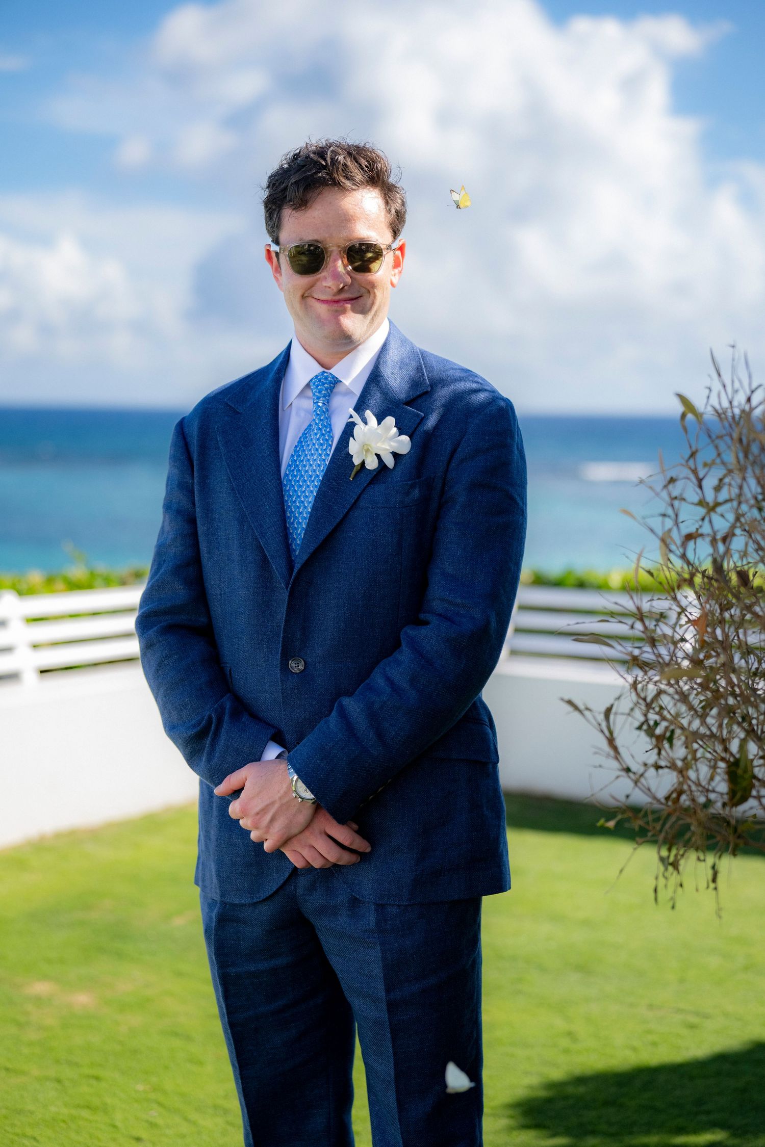 Groom in navy suit and blue tie stands against tropical ocean backdrop.