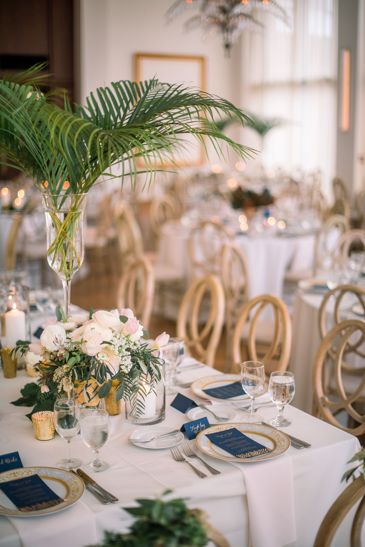 Elegant wedding reception tables decorated with palm fronds, navy blue accents, and gold details on crisp white tablecloths.