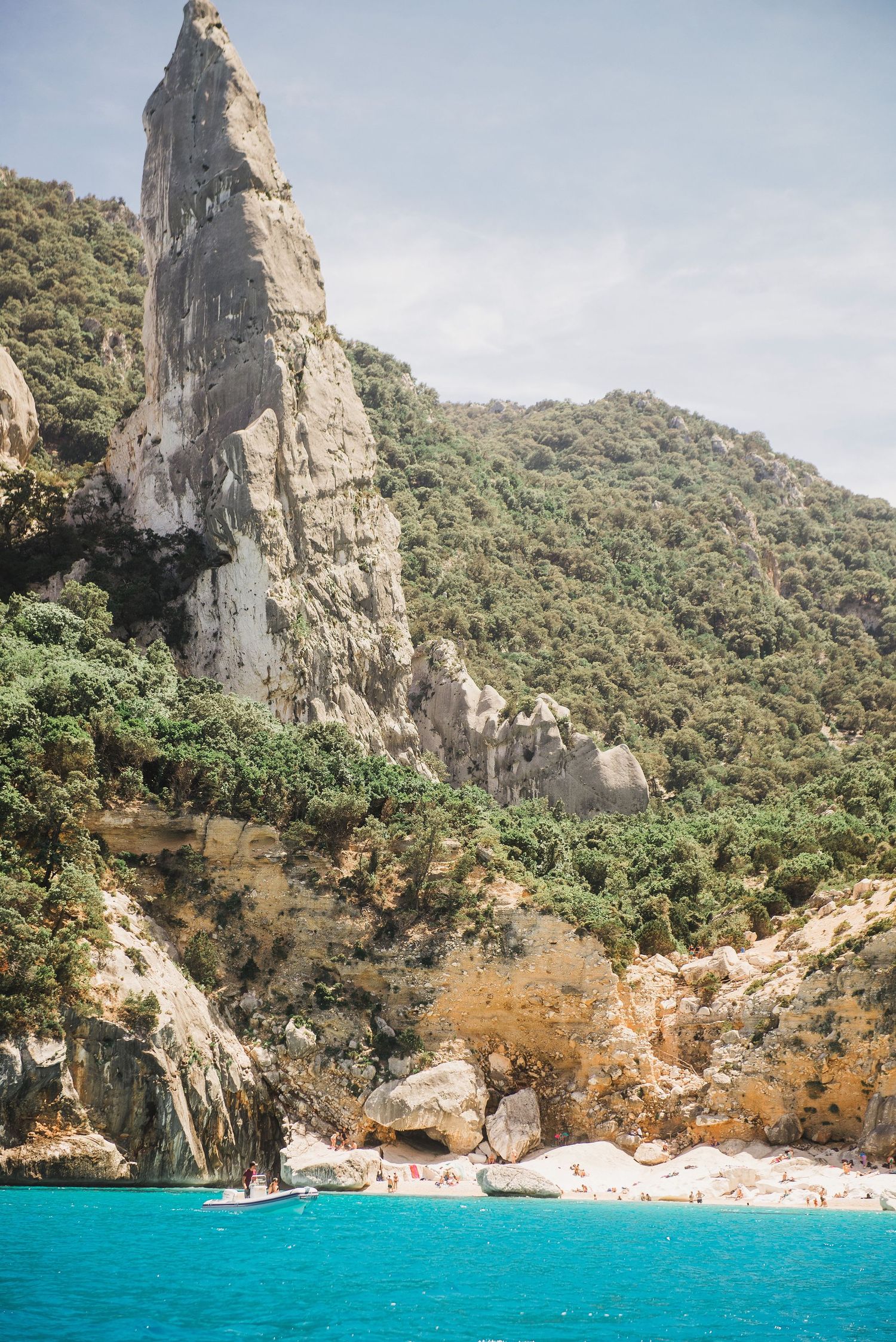 Dramatic limestone cliff face towers over a secluded turquoise beach cove with boats anchored along the Mediterranean coast.