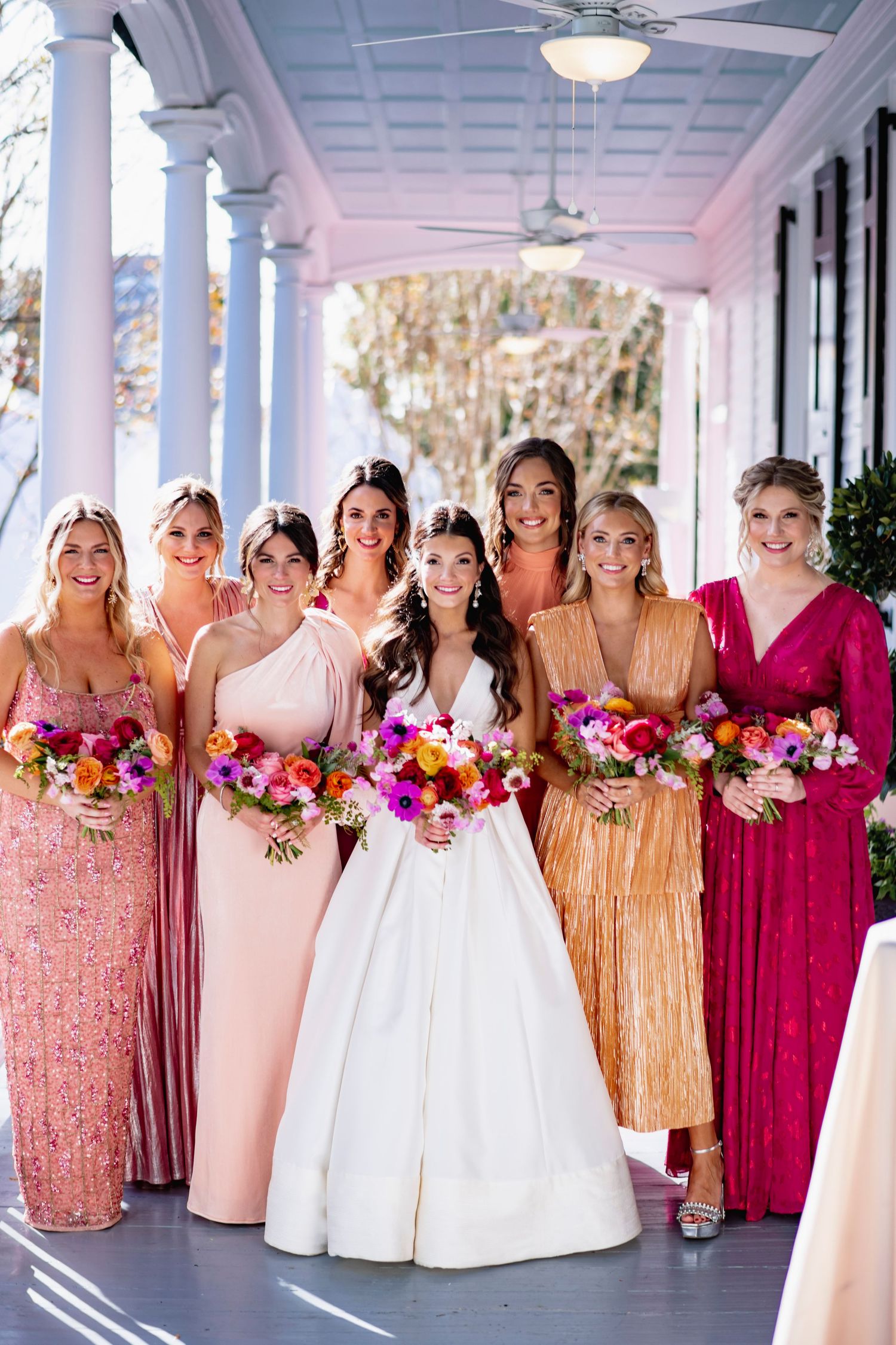 A bridal party in pink, blush and gold sequin dresses poses together on a white columned porch.