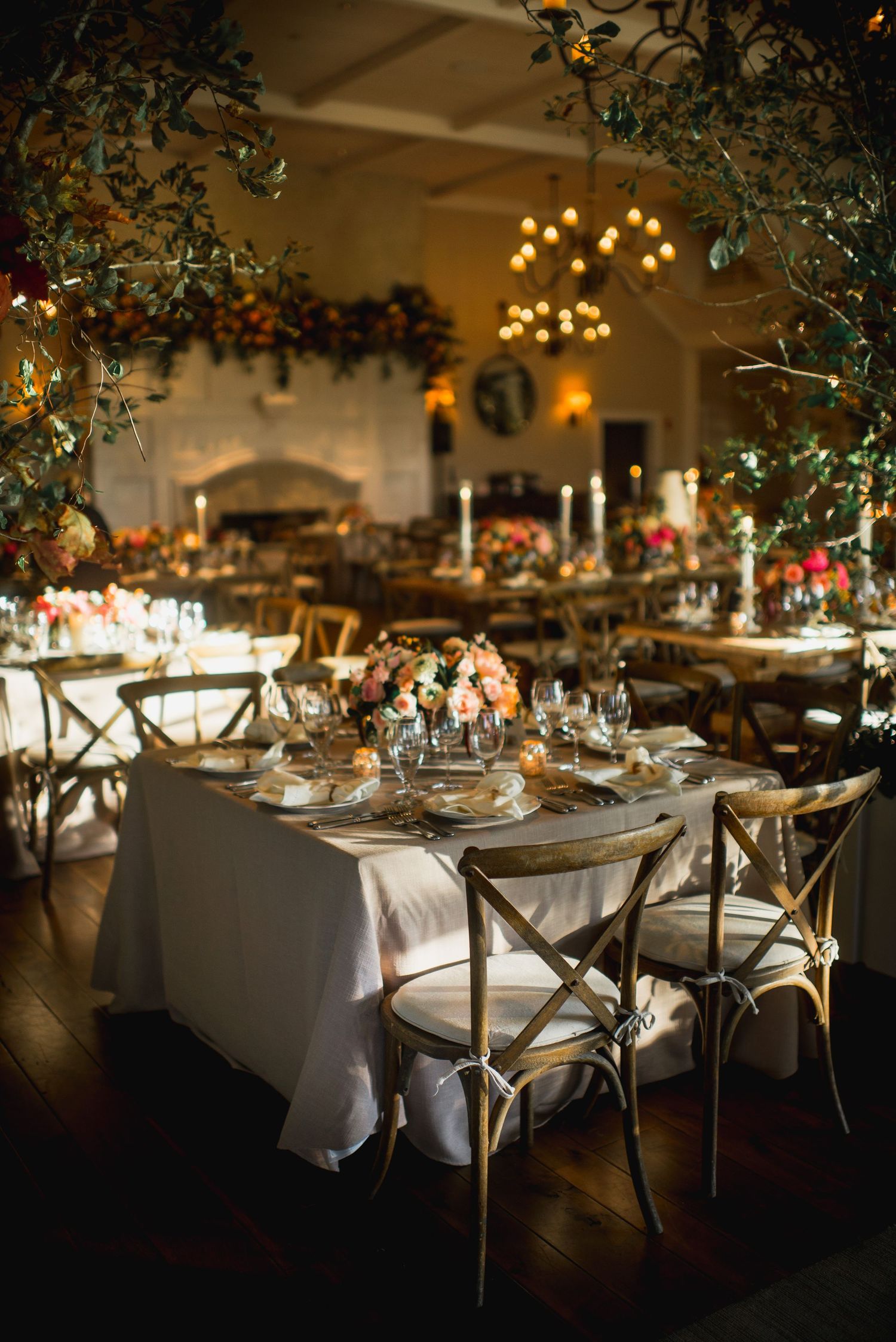 Elegant wedding reception tables with white linens, wooden cross-back chairs, and hanging floral arrangements in warm lighting.
