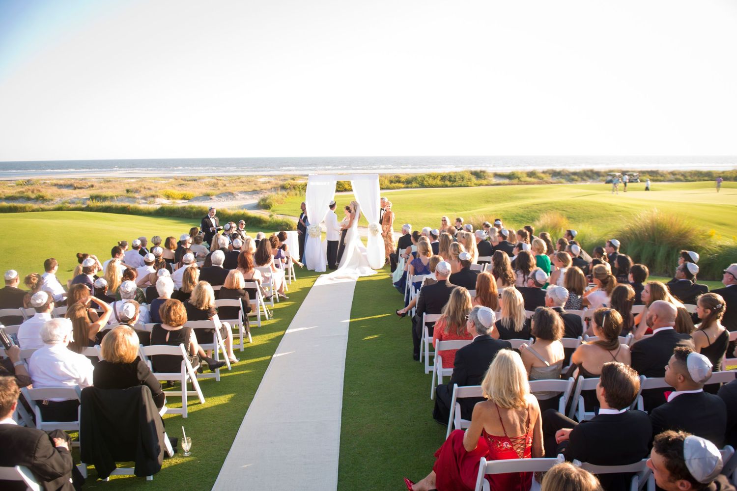 Wedding ceremony guests seated in white chairs overlooking a scenic coastal landscape at sunset.