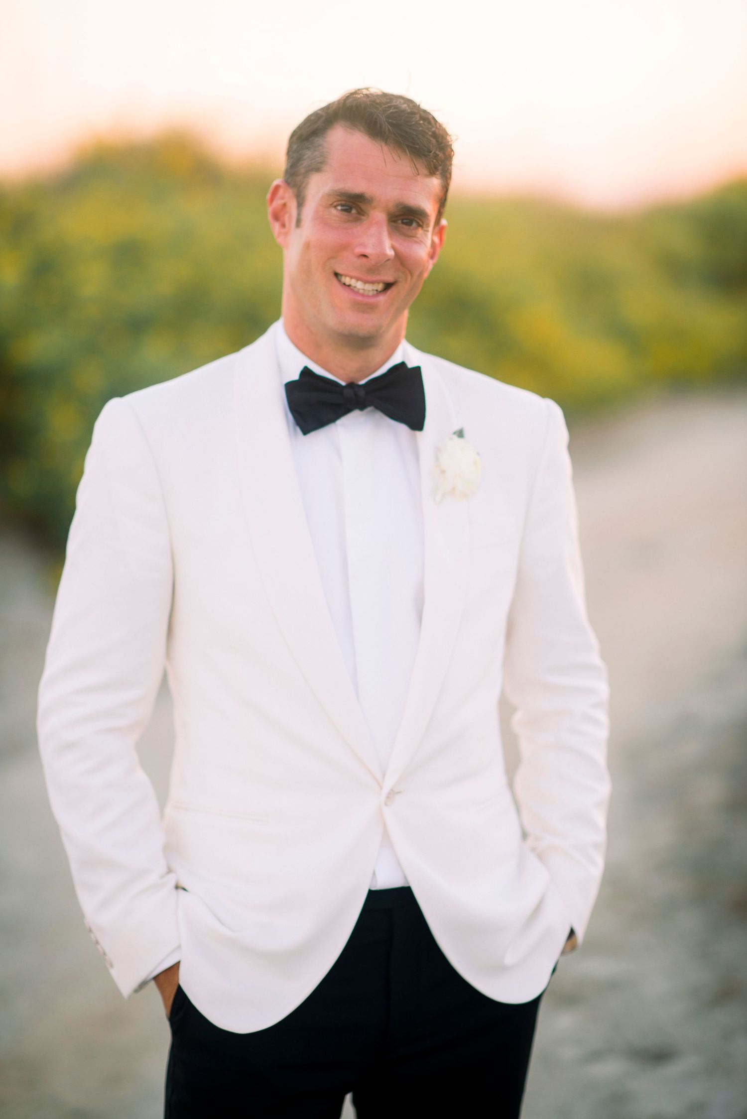A person wearing a white dinner jacket with black bow tie smiles while standing outdoors at dusk.