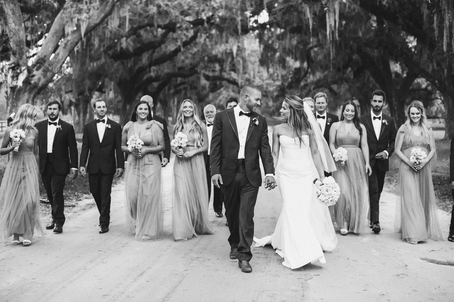 Wedding party members walk in a line across a grassy lawn during an outdoor celebration.