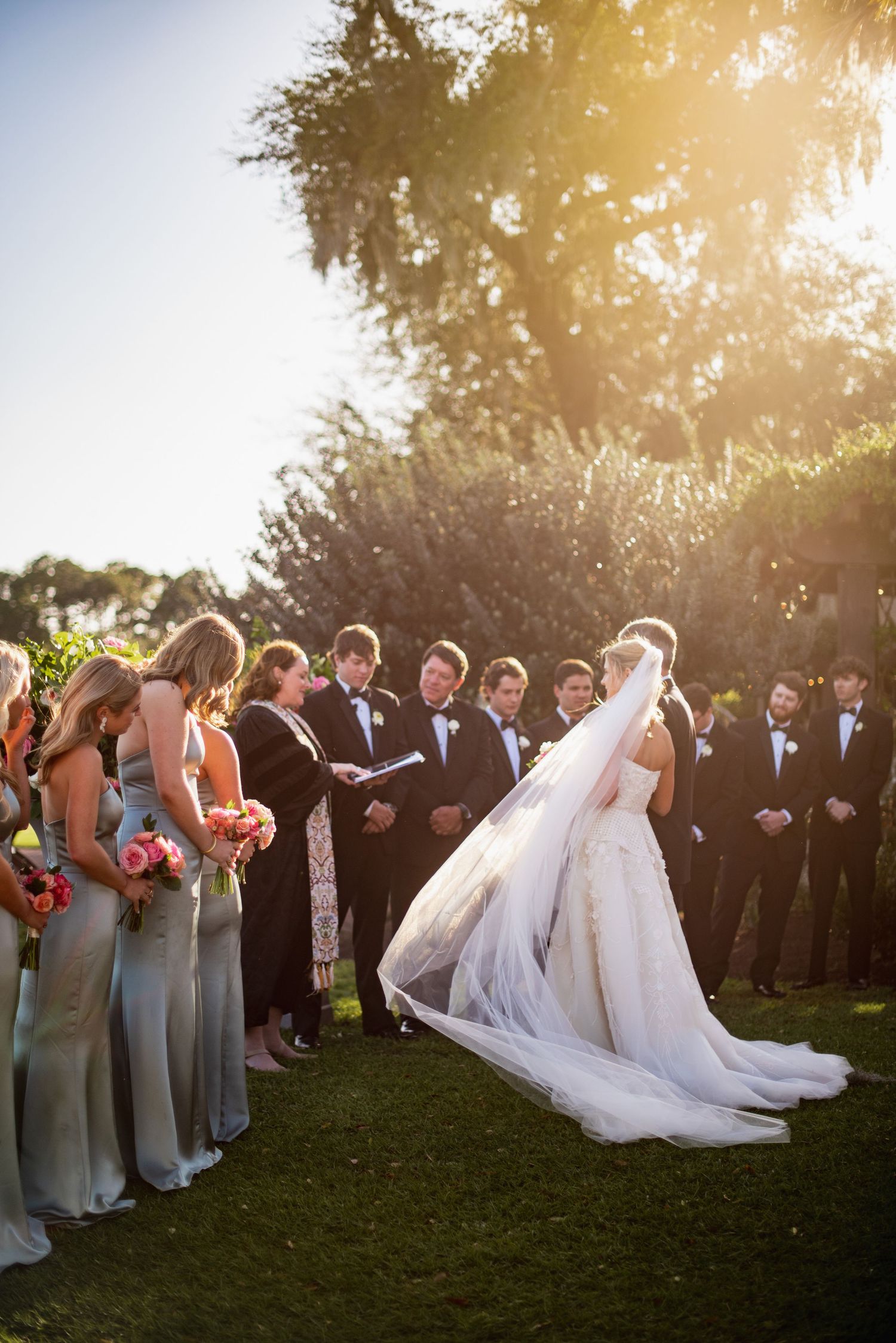 Wedding party in formal attire poses outdoors at sunset with bridesmaids in gray dresses and groomsmen in dark suits.