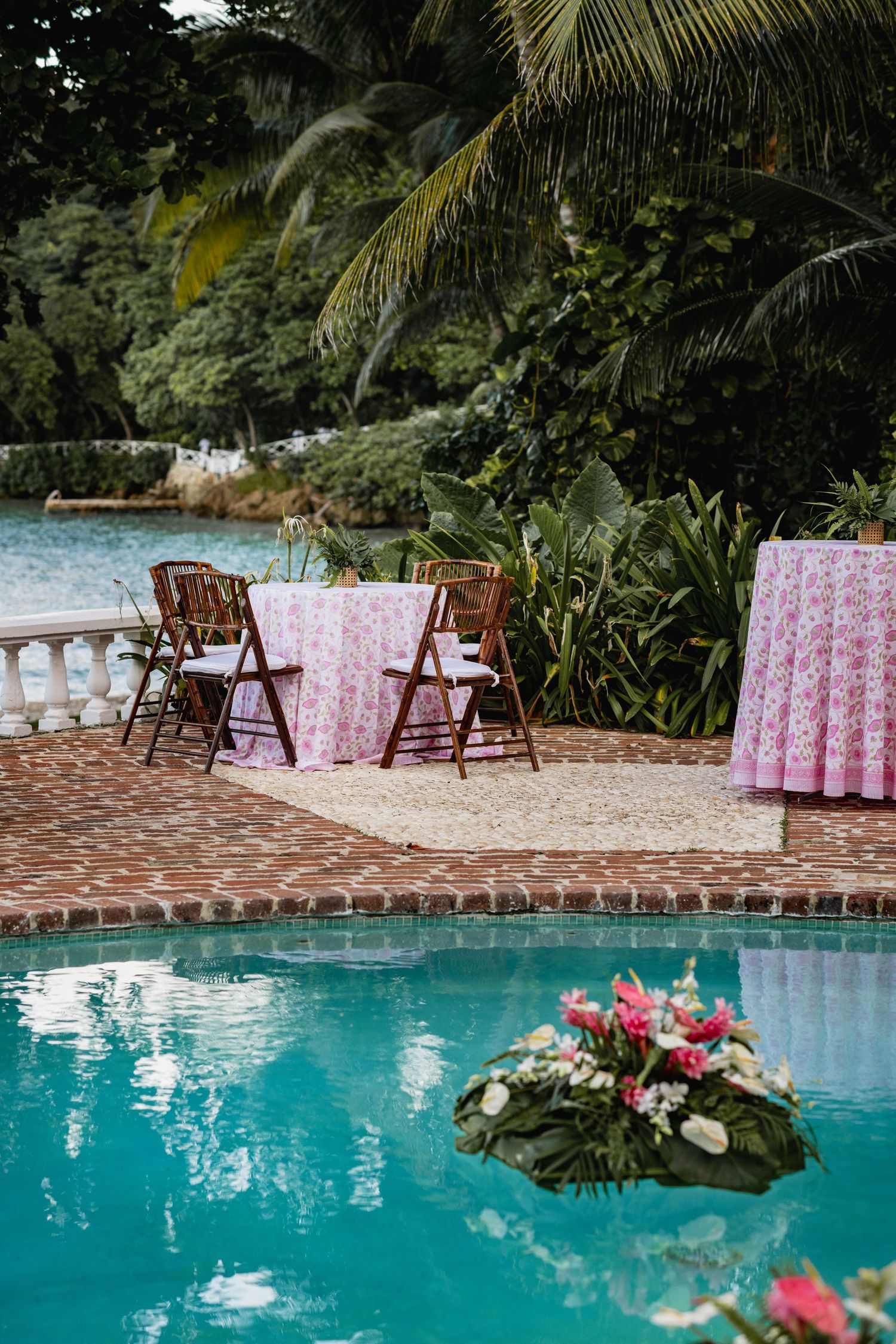 Wooden chairs with pink floral tablecloths arranged around a turquoise swimming pool with tropical palm trees in background.