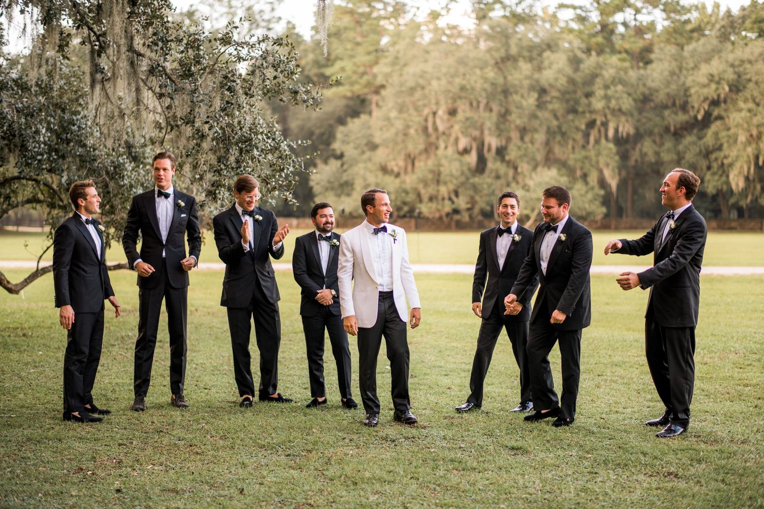 Groomsmen in formal attire laughing together on a grassy lawn with Spanish moss and trees in the background.