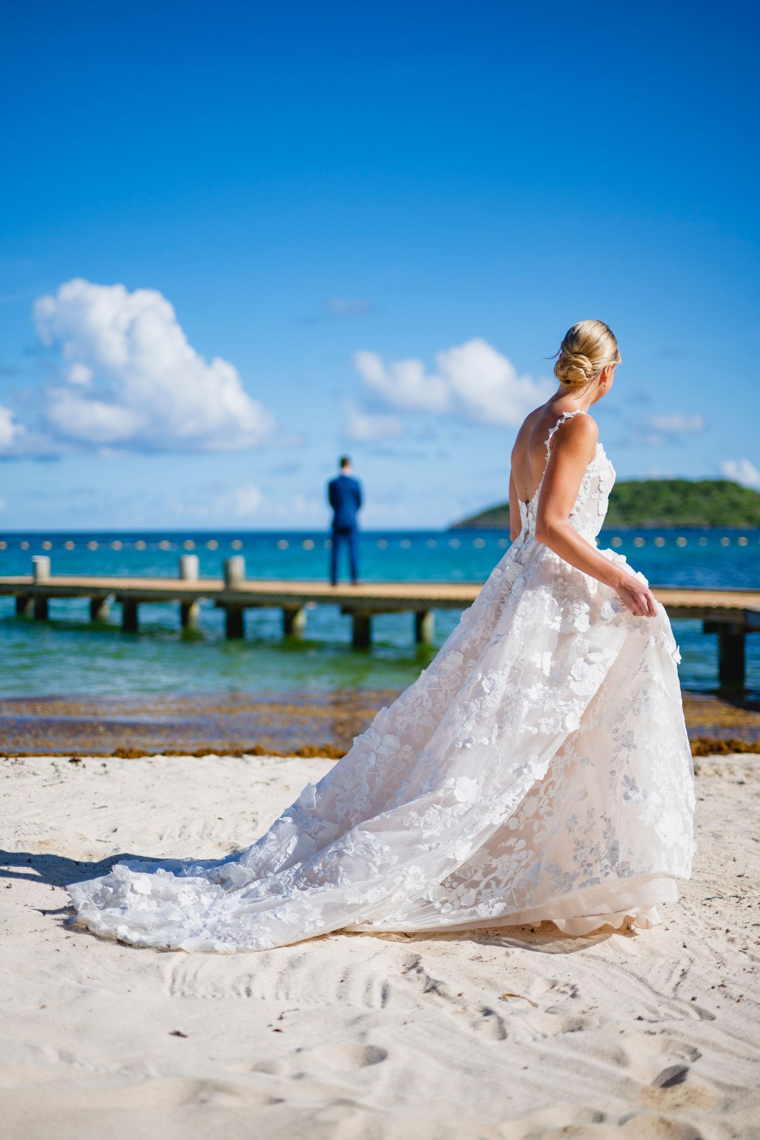 White lace wedding dress flowing across sandy beach with ocean and wooden pier in background.