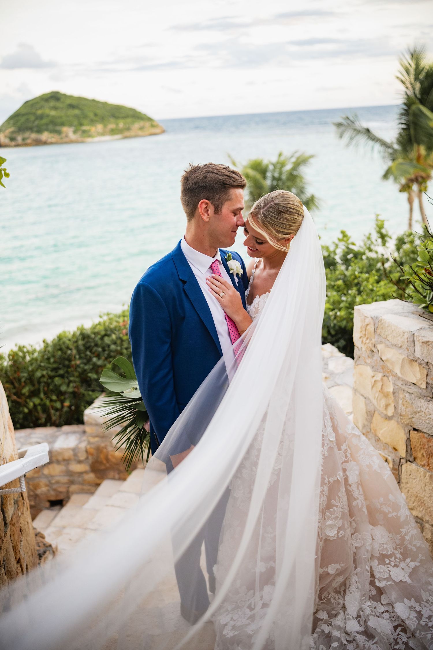 Wedding couple embraces against an ocean backdrop with flowing veil and blue suit.