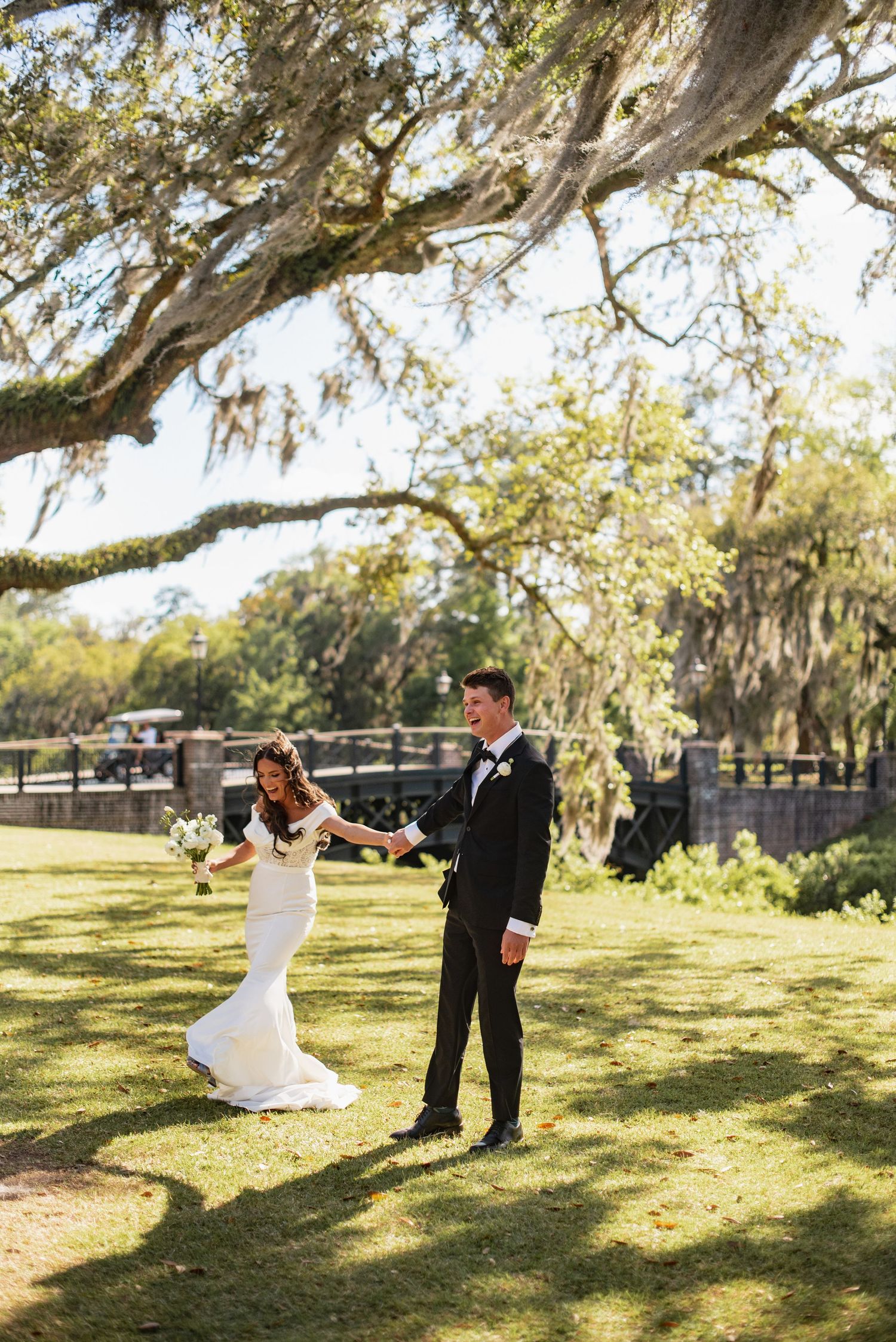 A couple in wedding attire playfully poses under Spanish moss-draped trees in a sunlit outdoor setting.
