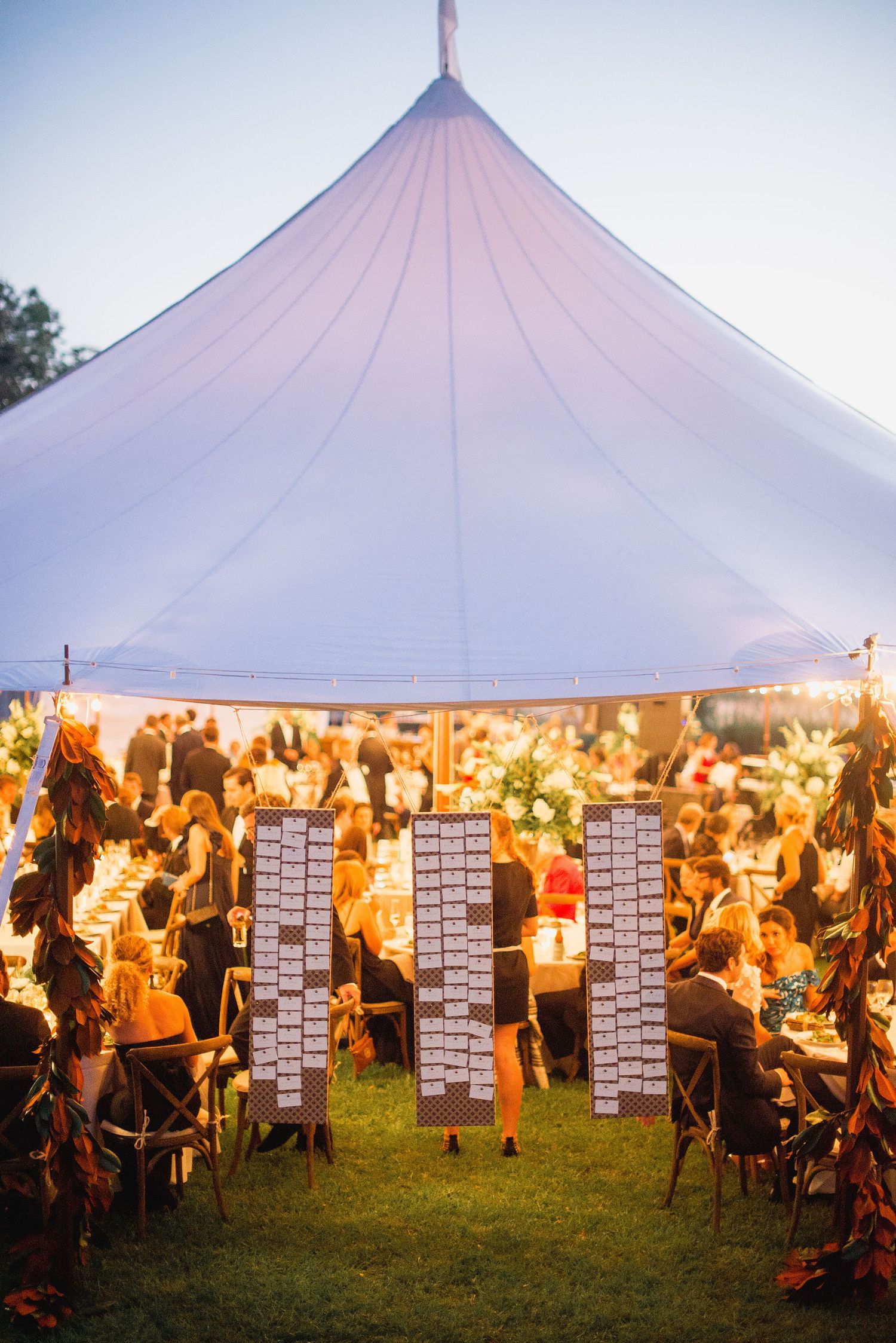 Large white wedding tent illuminated at dusk with guests seated at tables beneath glowing string lights.
