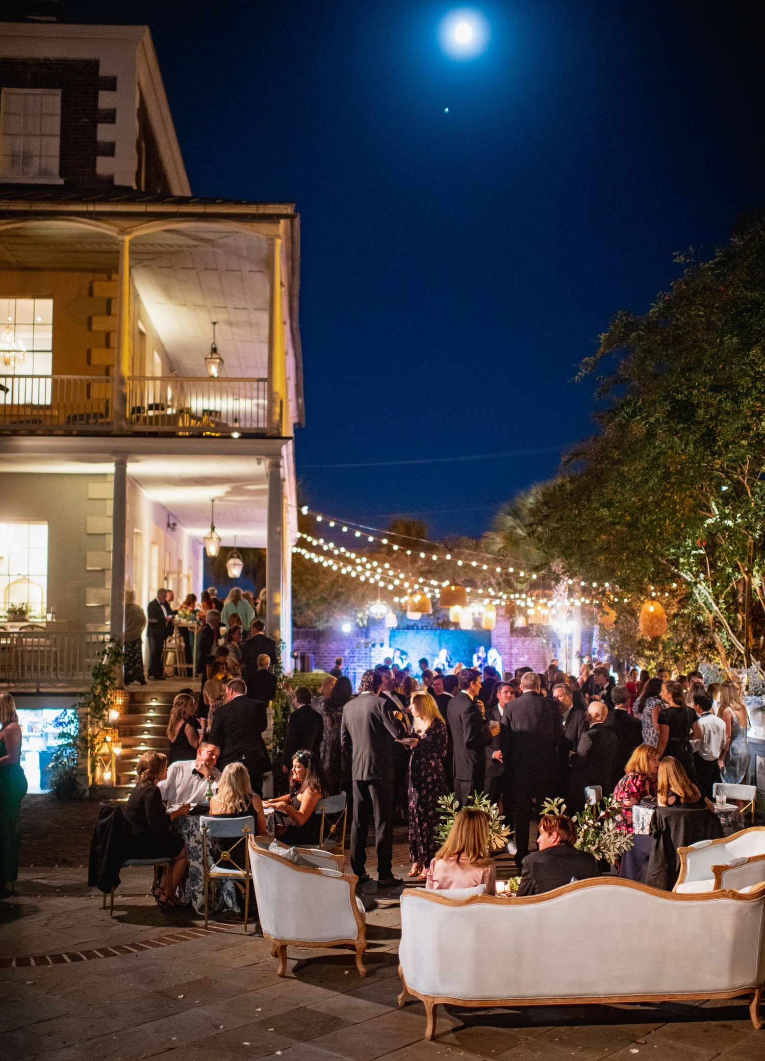 Elegant outdoor evening party with string lights and a historic two-story house featuring a crowded patio and balcony.