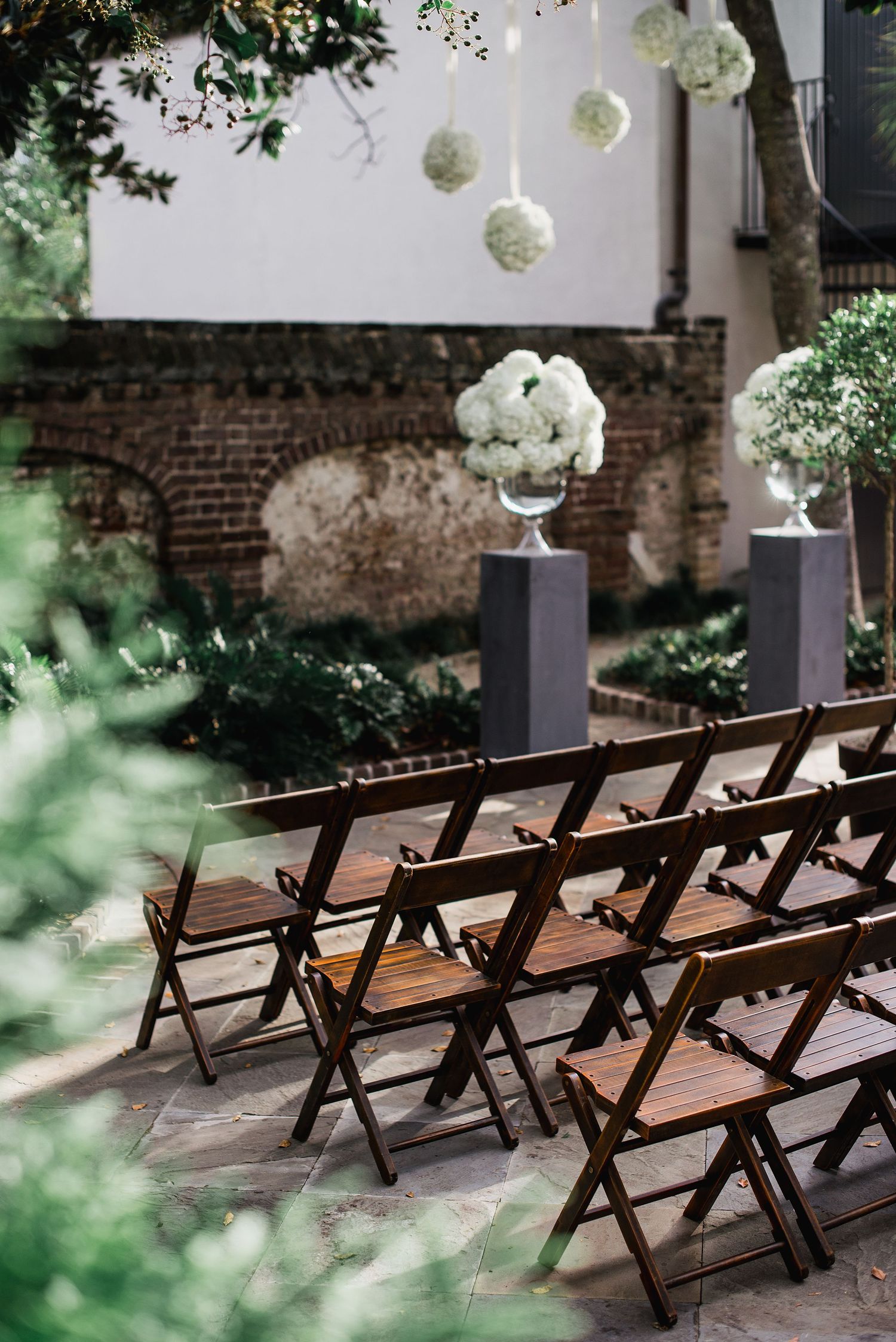 Elegant outdoor wedding ceremony setup with wooden folding chairs and white floral arrangements on gray pedestals.