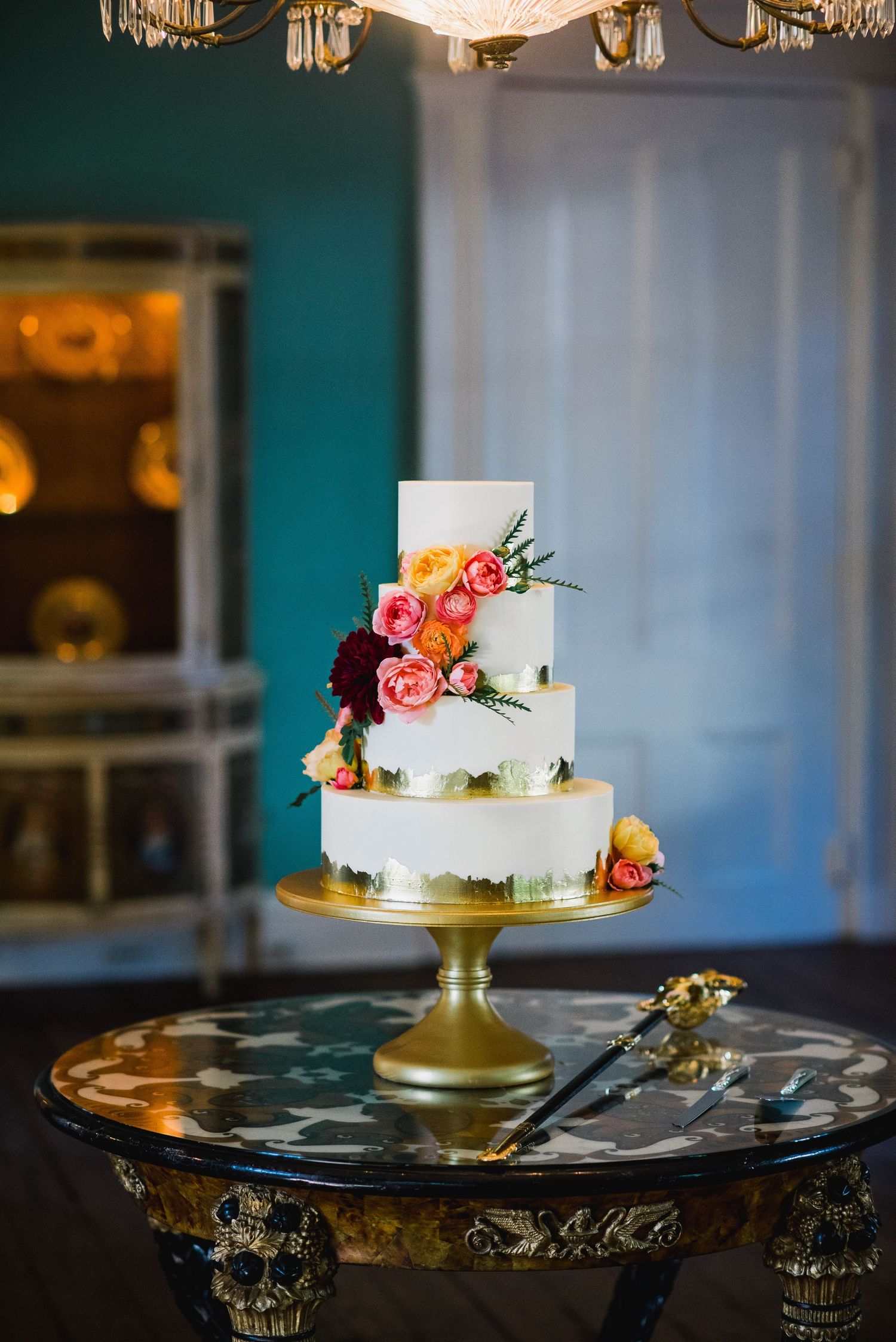White wedding cake with coral flowers and greenery displayed on an ornate gold cake stand with elegant lighting.