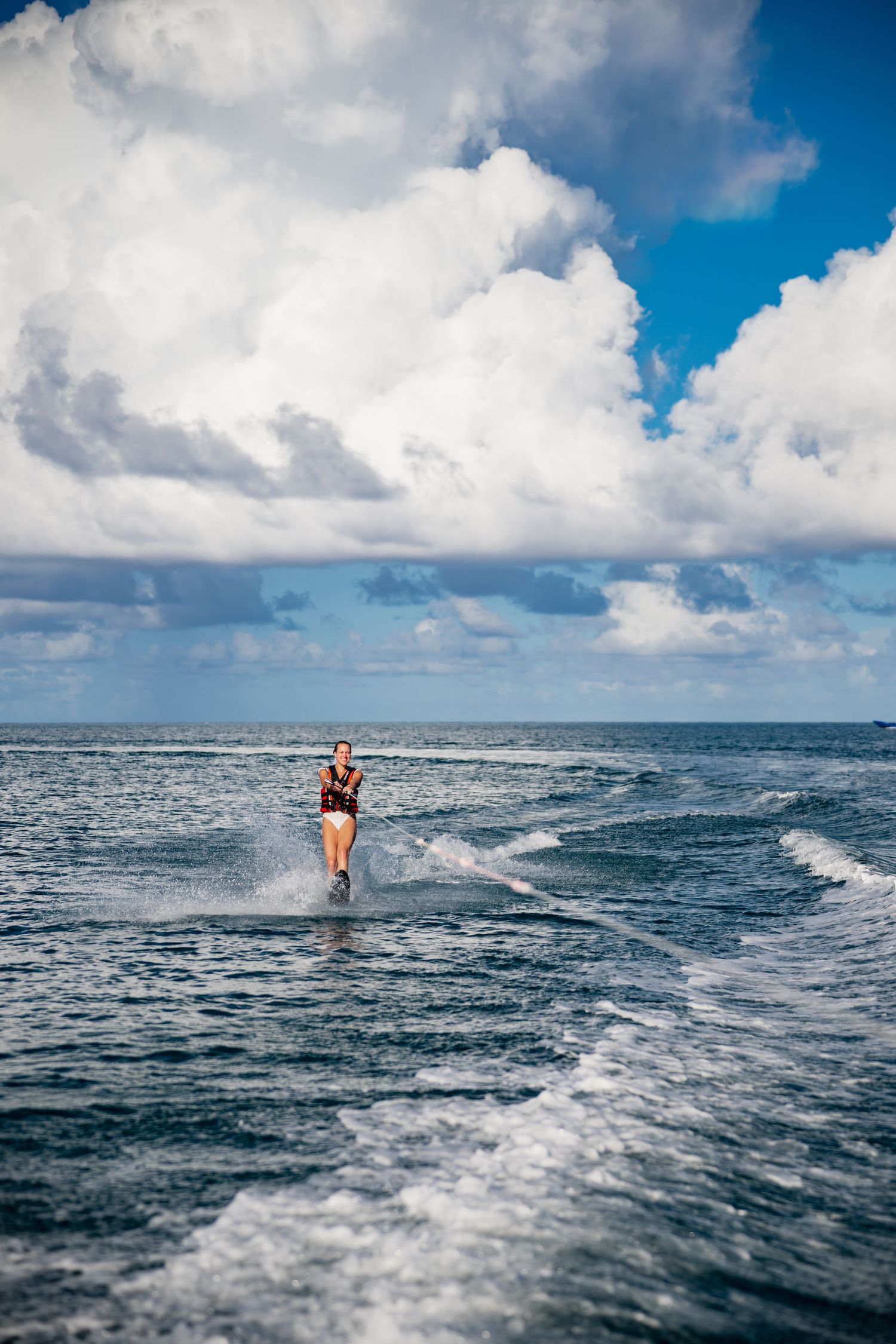 A person waterskiing across blue ocean waters under dramatic white clouds and bright blue sky.