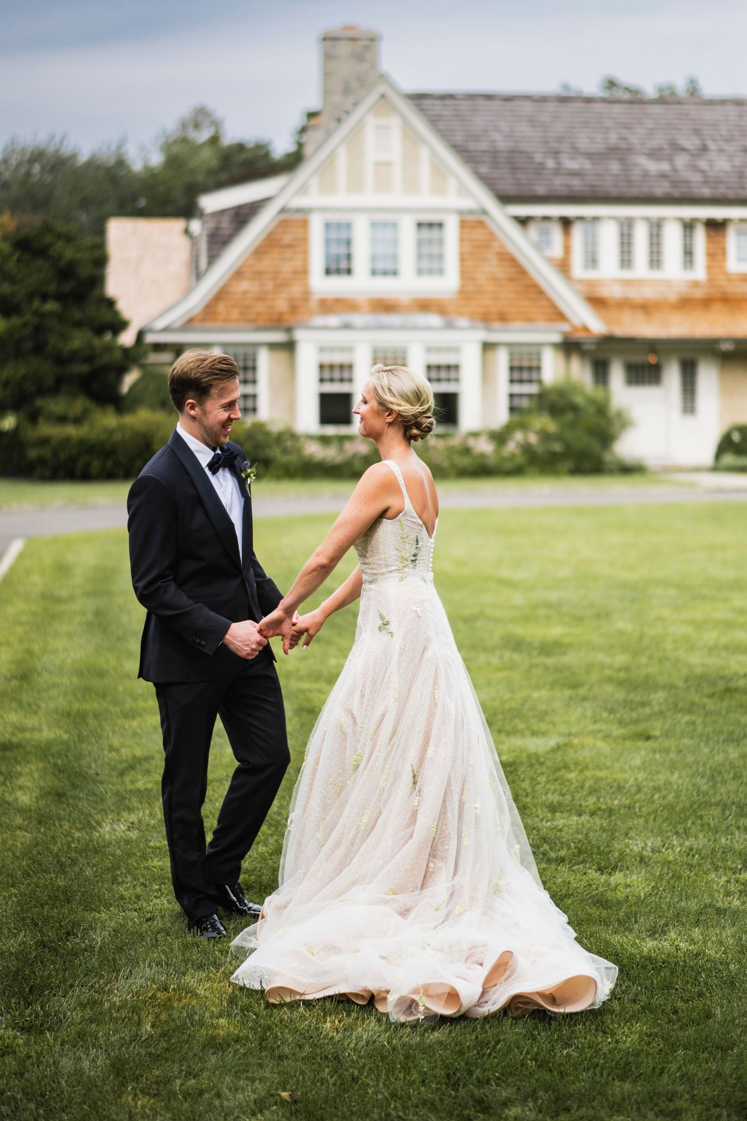 Newlyweds in formal attire playfully interact on a grassy lawn in front of historic buildings.