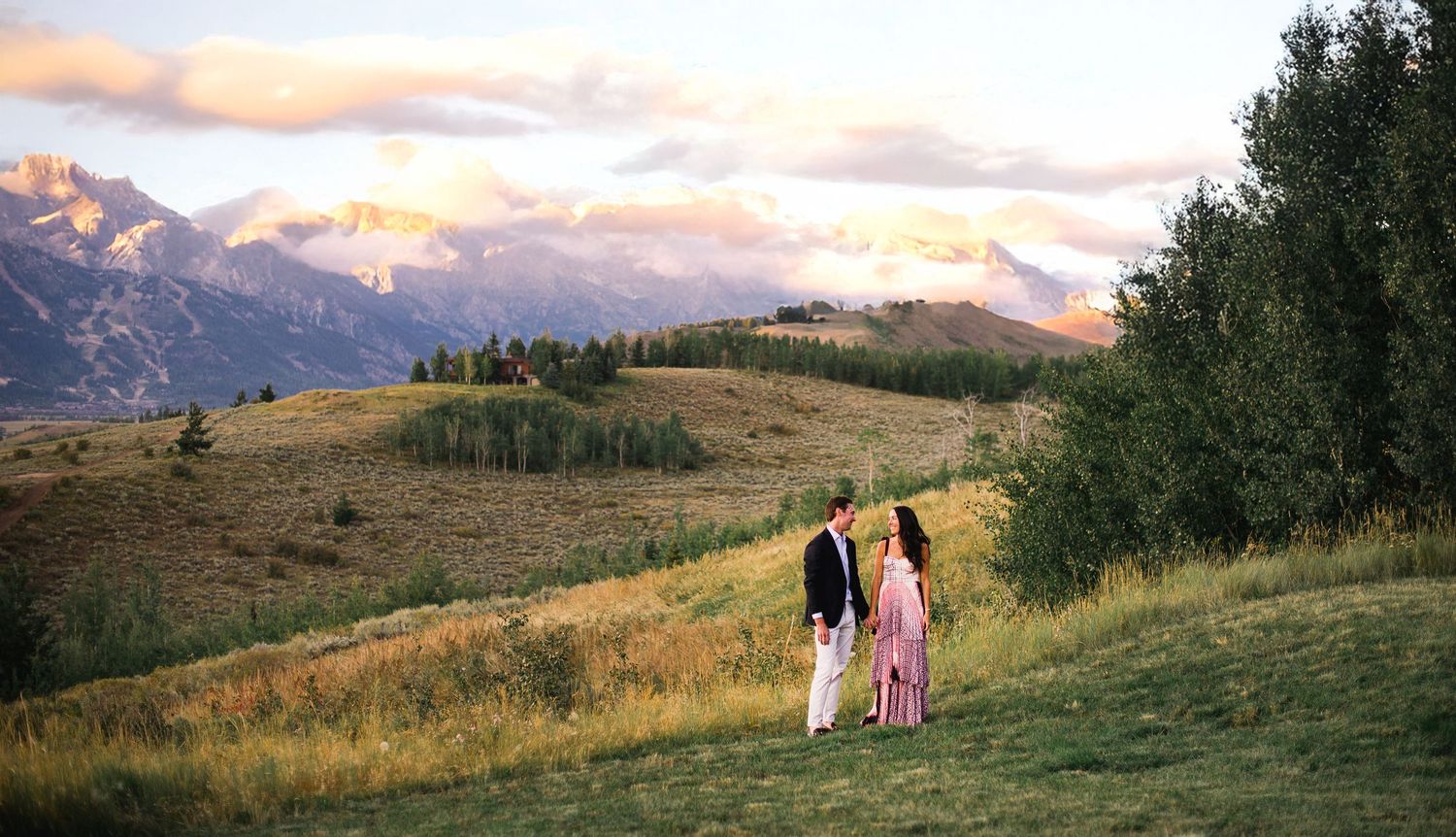 Two people walking on a grassy mountain slope at sunset with majestic mountain peaks and forests in the background.