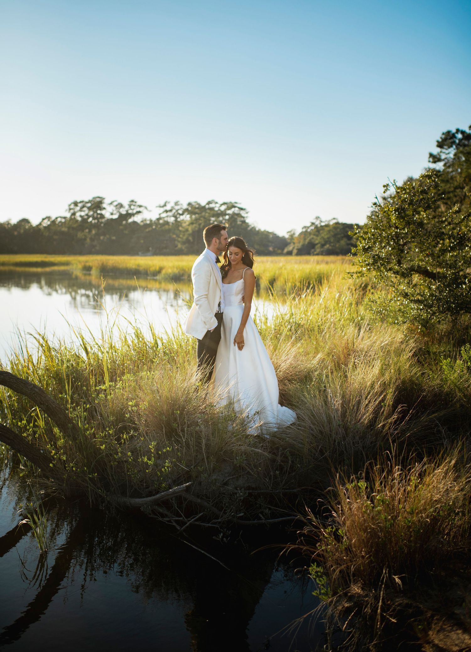 A romantic sunset silhouette of a couple in wedding attire standing by a serene marsh with golden light streaming through.