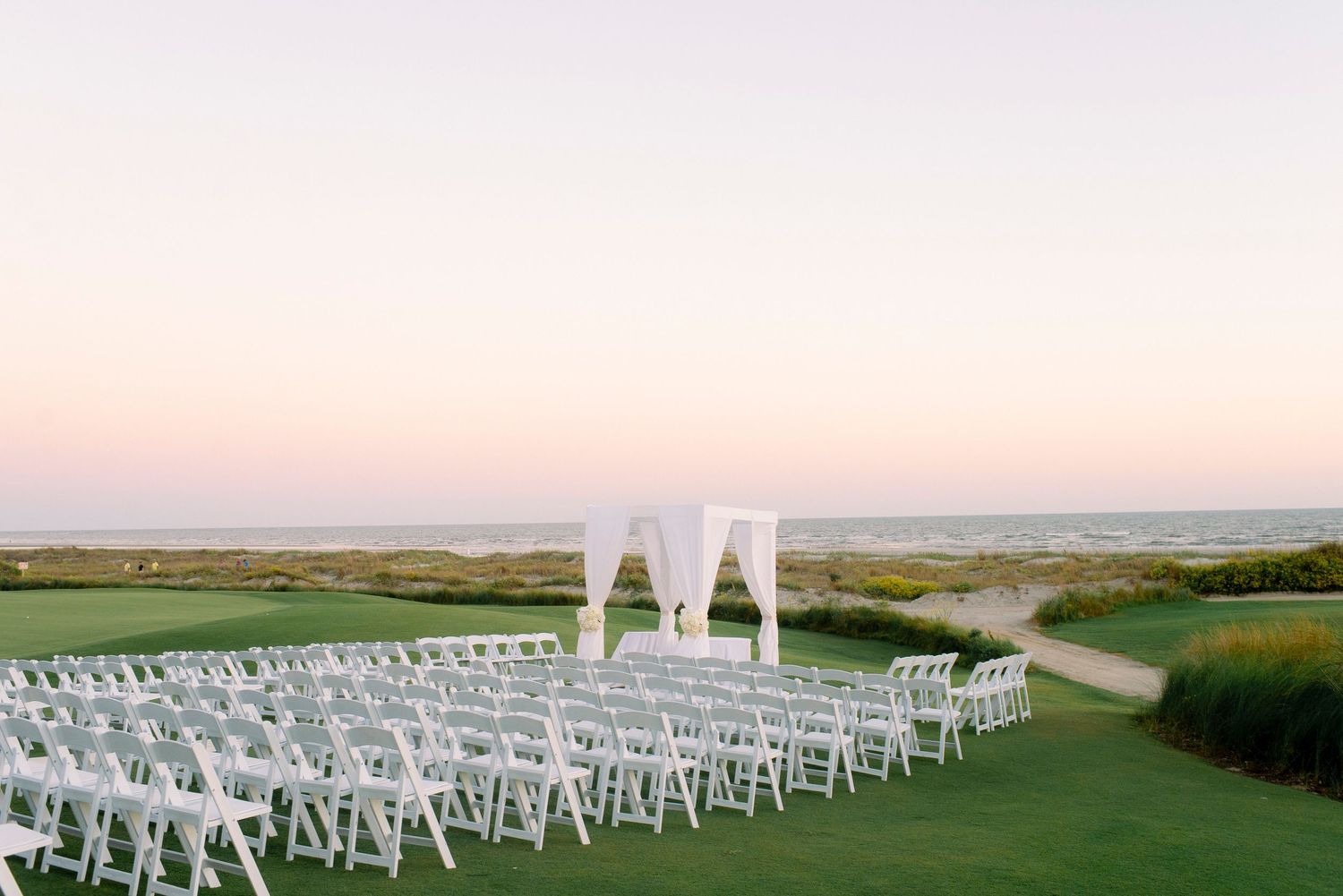 White chairs arranged for an outdoor wedding ceremony on a lush golf course with a white arch at sunset.