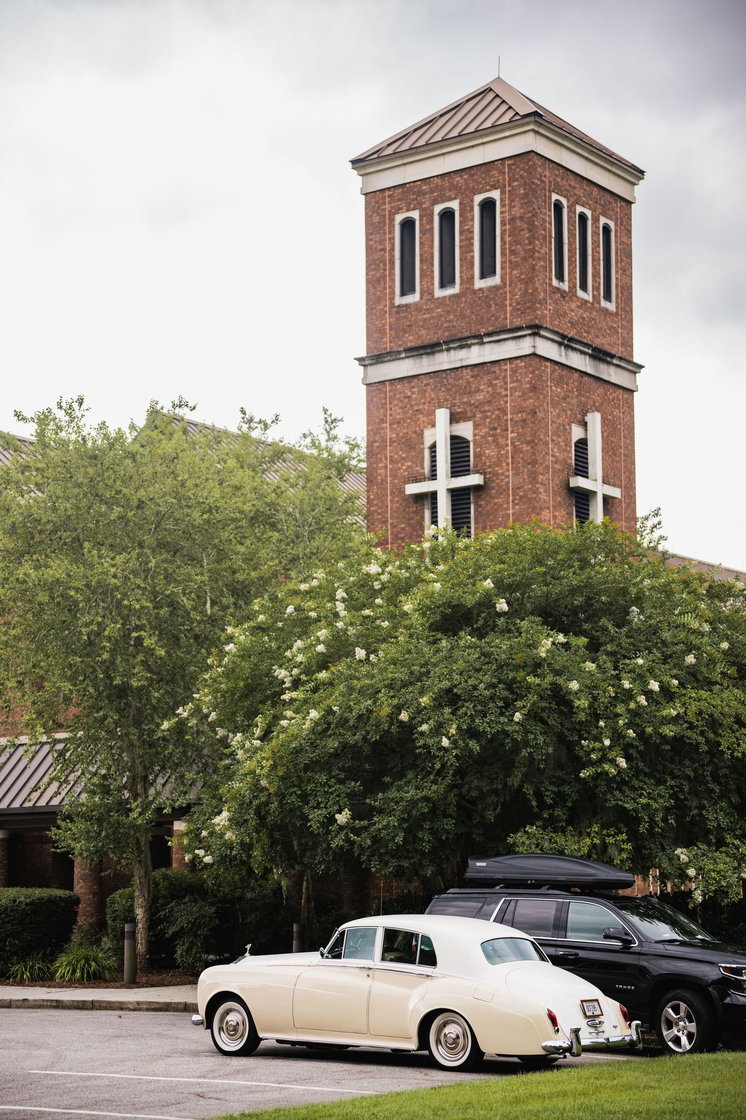 Classic vintage cars parked outside a red brick church with blooming trees in the foreground.