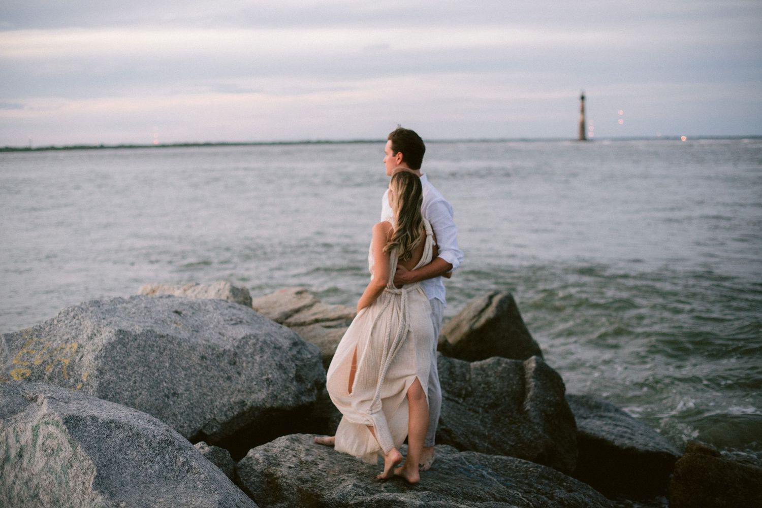 A person in a flowing white dress stands on coastal rocks overlooking a lighthouse in the distance.