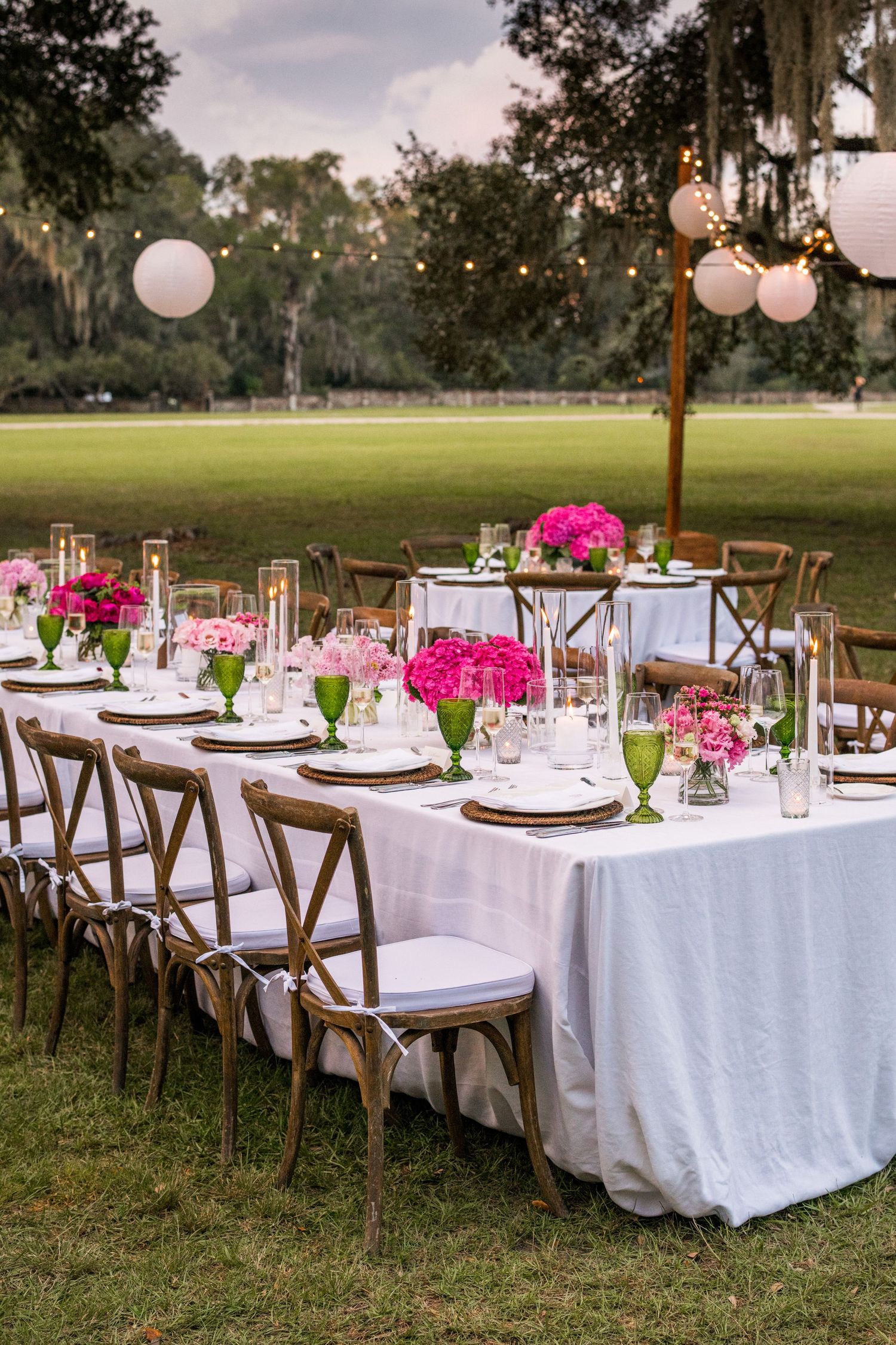 Elegant outdoor wedding reception table with white linens, wooden chairs, pink floral centerpieces and hanging lanterns at dusk.
